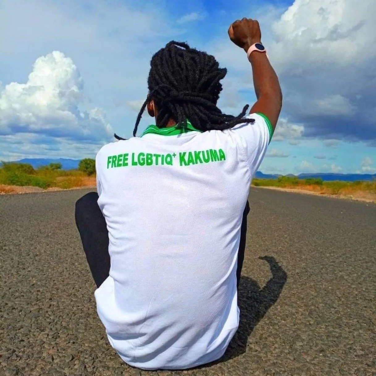 A person with dreadlocks sits on an empty road, facing away, wearing a white T-shirt with green text that reads 'FREE LGBTIQ+ KAKUMA.' They are raising a fist in the air, with a scenic landscape of blue sky and distant mountains in the background.
