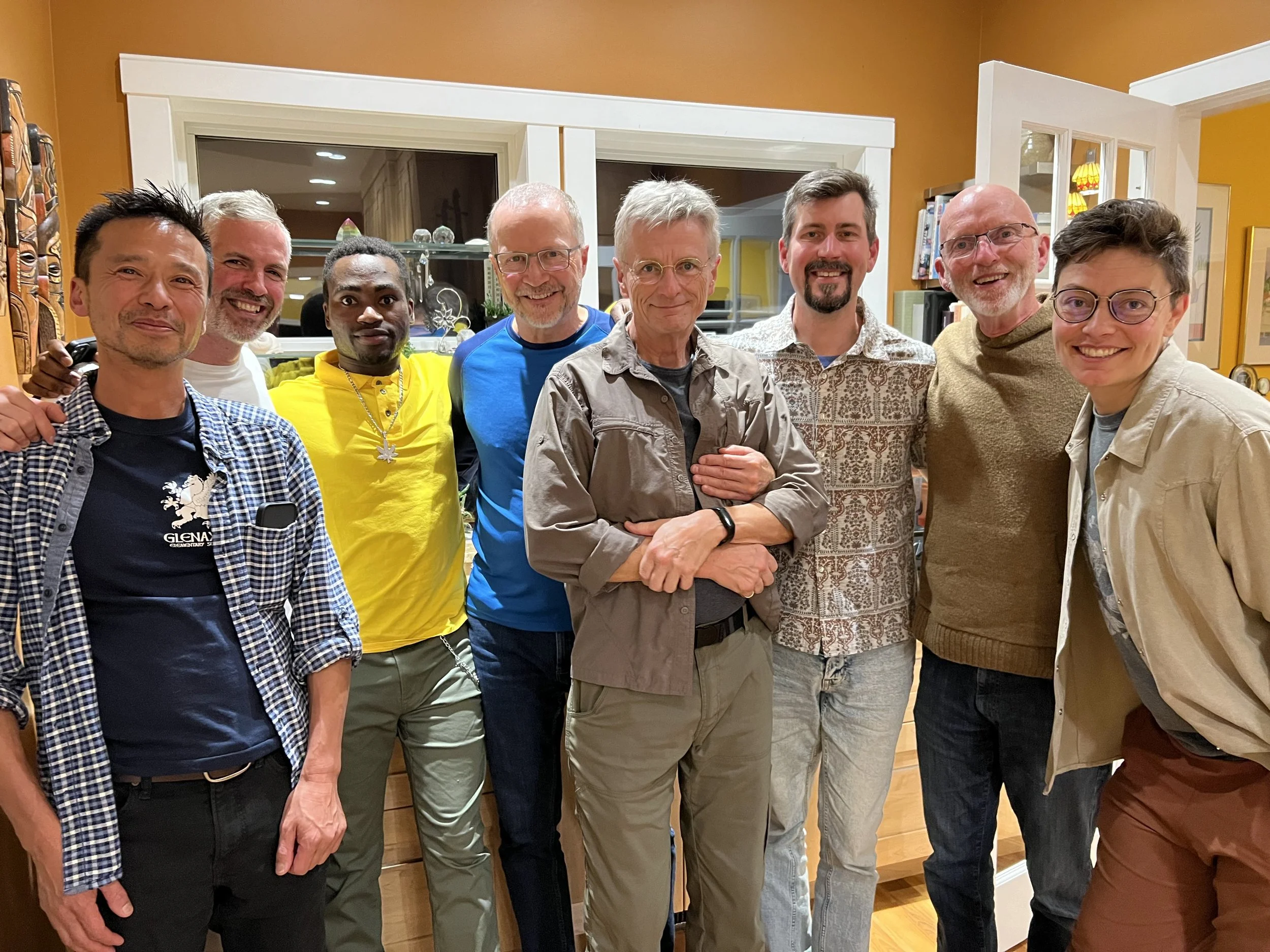 A group of nine men posing together indoors, smiling, in front of a yellow wall with kitchen and decorative items visible in the background.