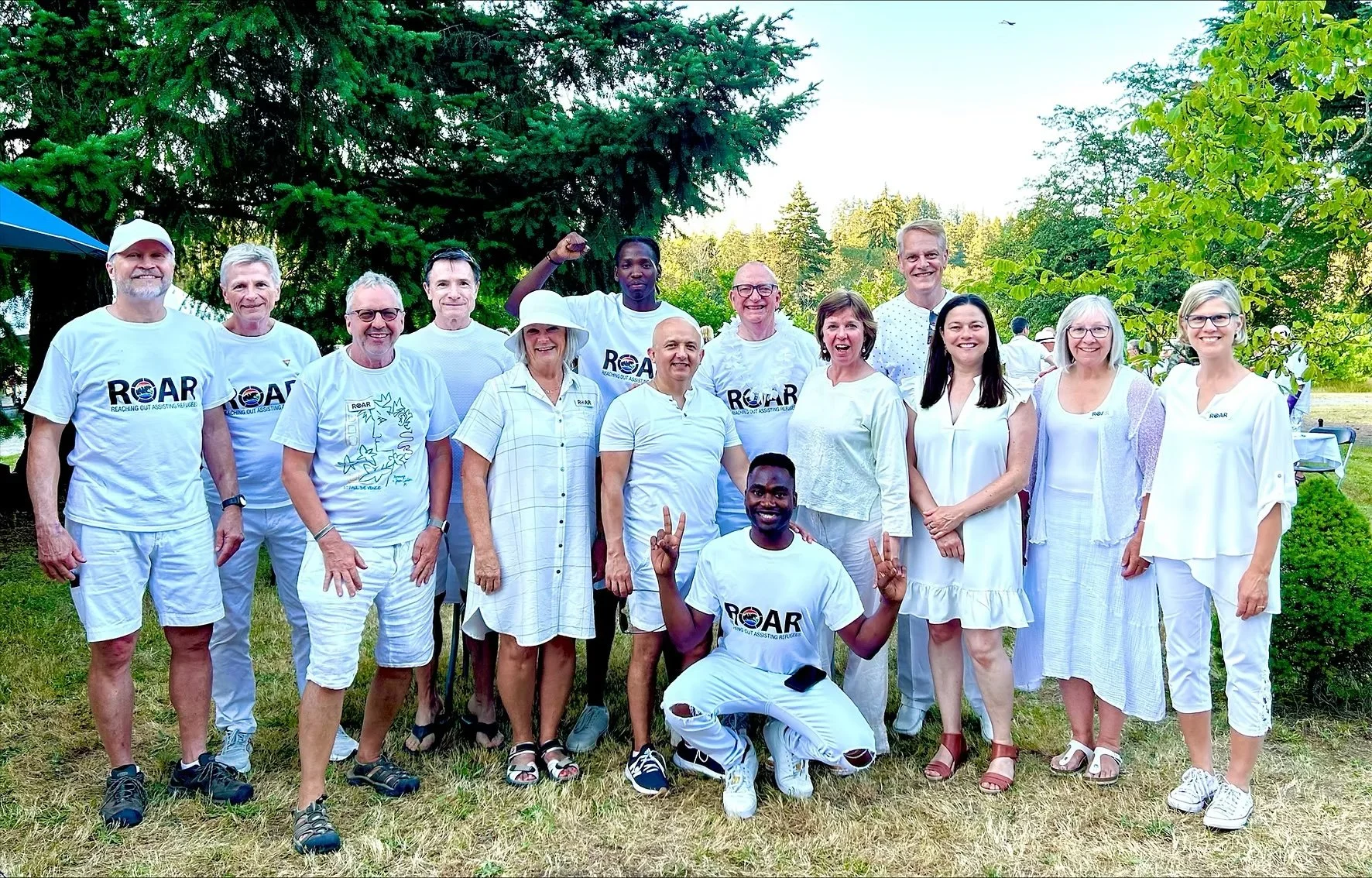 Group of people outdoors, some wearing white shirts with 'ROAR' logo, standing on grass with trees in the background, smiling and posing for the photo.