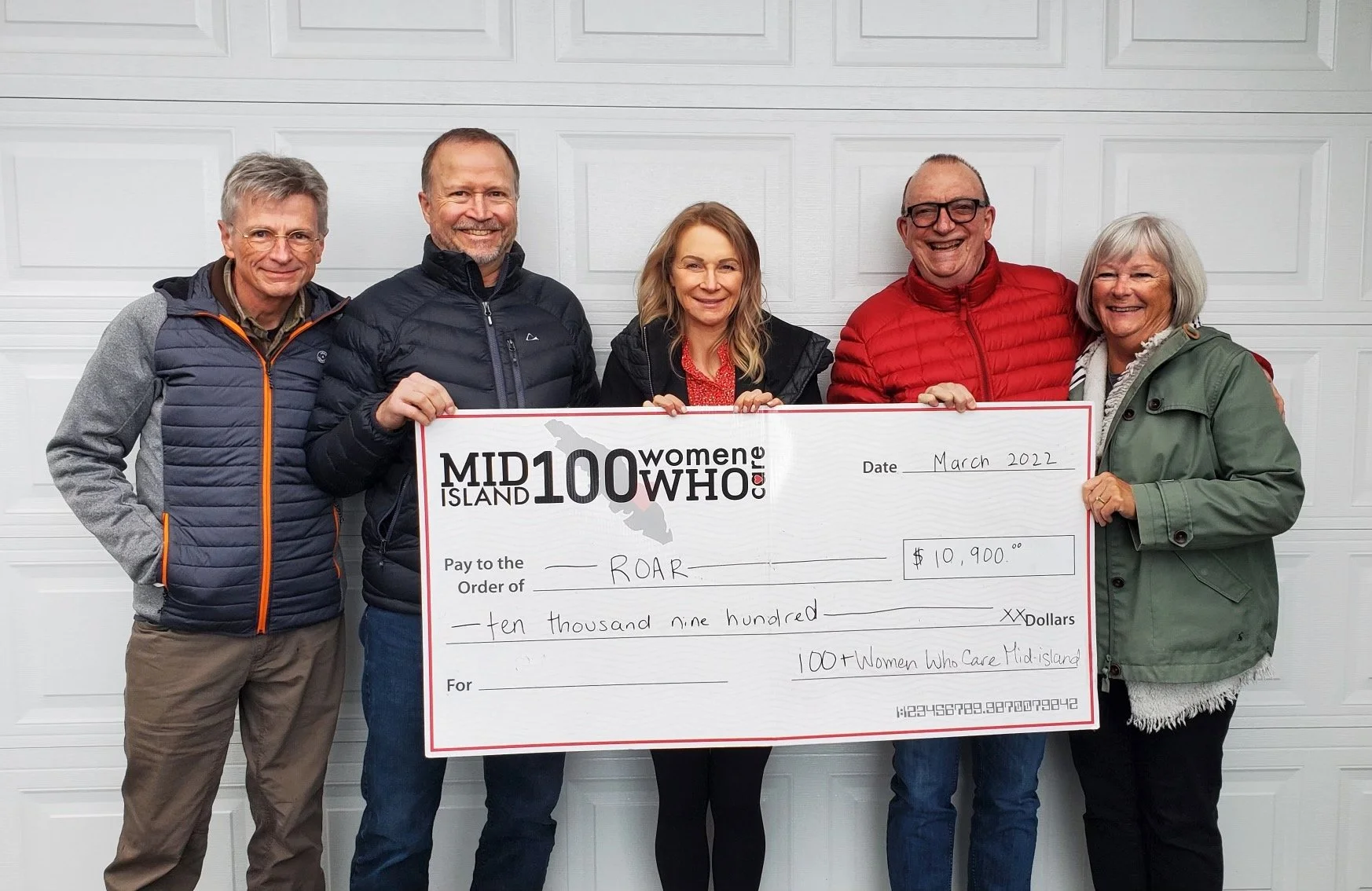 Five people standing in front of a white garage door, holding a large check made out to ROAR for ten thousand nine hundred dollars, dated March 2022, with the description '100 Women Who Care Mid-Island.'