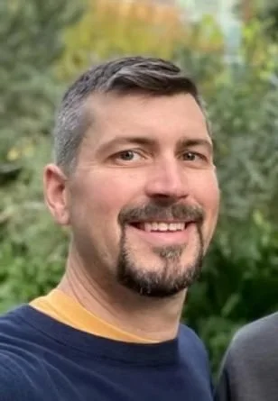 A smiling man with short dark hair and a beard, outdoors with trees and greenery in the background.