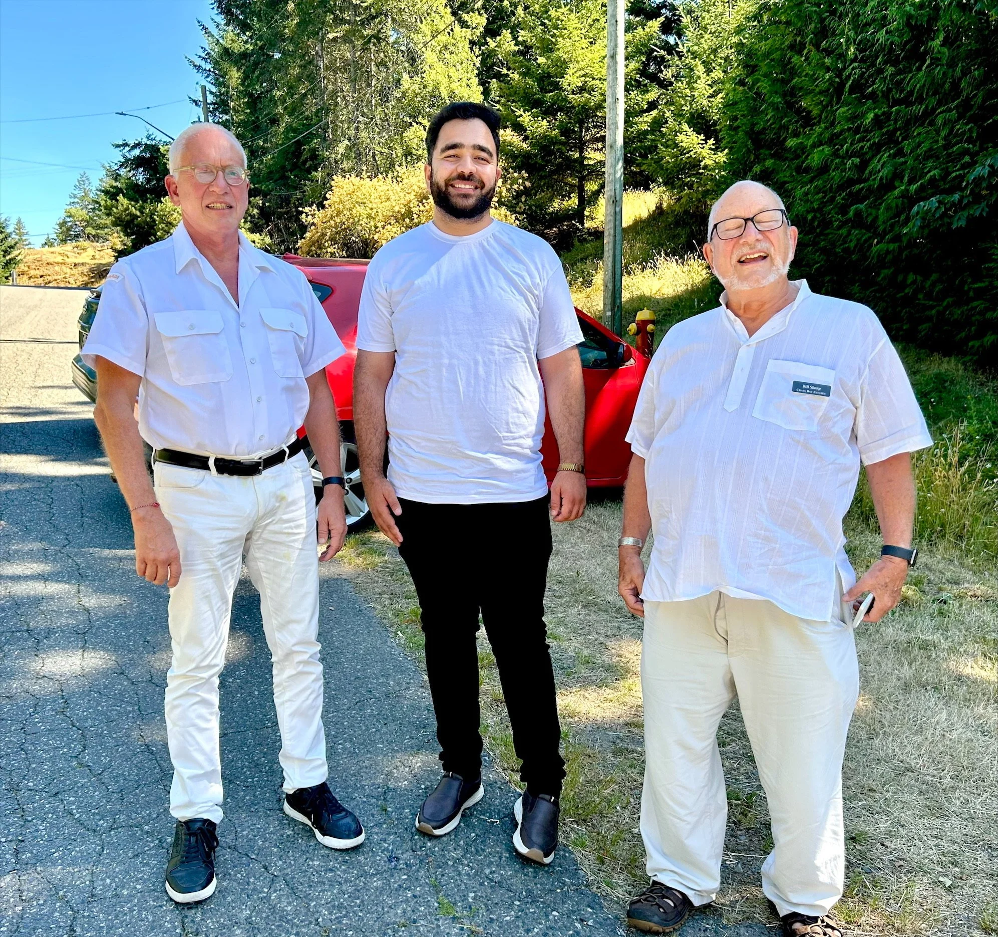 Three men standing outdoors on a paved road beside a red car, with green trees and foliage in the background, sunny weather, and all three men wearing casual white clothing and smiling.