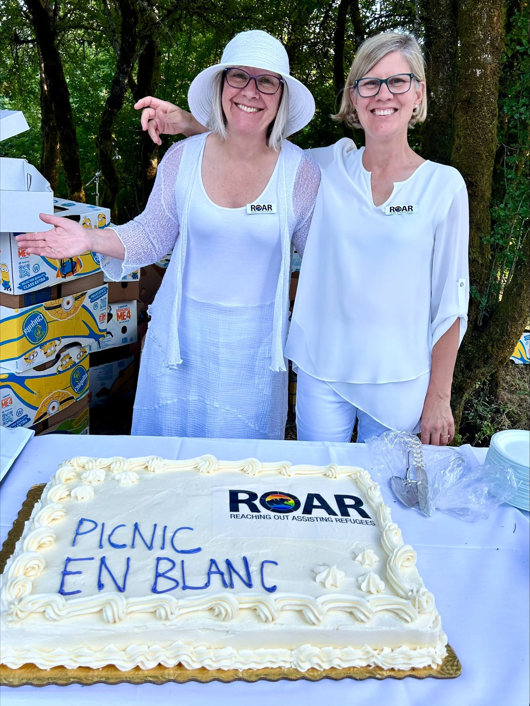 Two women in white shirts and glasses standing behind a table with a large white cake that has the ROAR logo and the words 'PICNIC EN BLANC' written on it. They are outdoors, surrounded by trees and boxes of food or supplies.