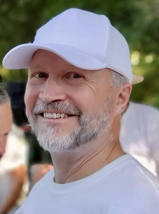 A smiling middle-aged man with a beard and mustache wearing a white baseball cap and white shirt, outdoors with a blurred background of greenery.