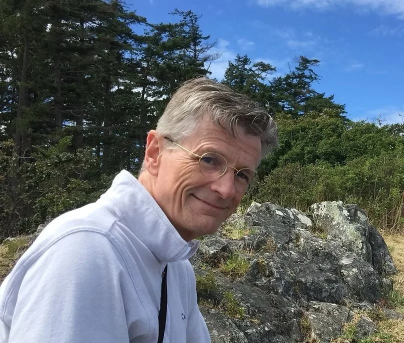 A man with glasses and gray hair sitting outdoors near rocks and trees, smiling at the camera.