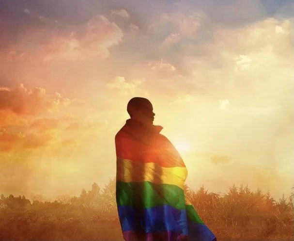 Person standing outdoors at sunset, wrapped in a rainbow pride flag.