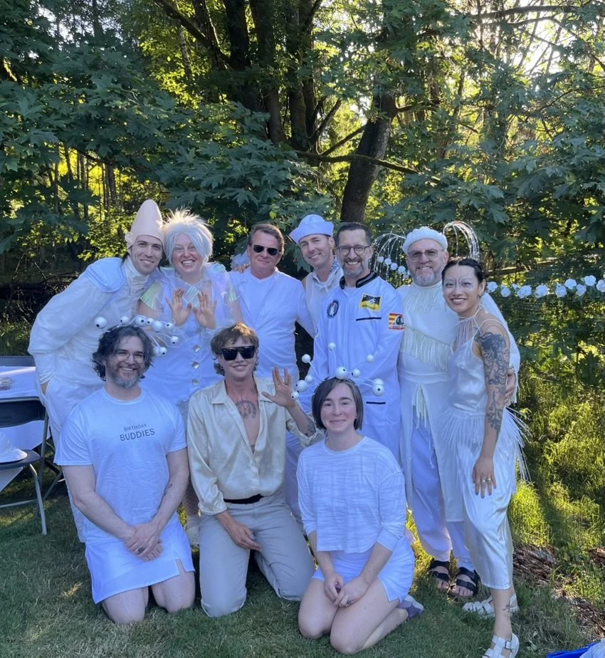 Group of people dressed in white costumes with creative and playful accessories, posing outdoors near trees, celebrating a special occasion.