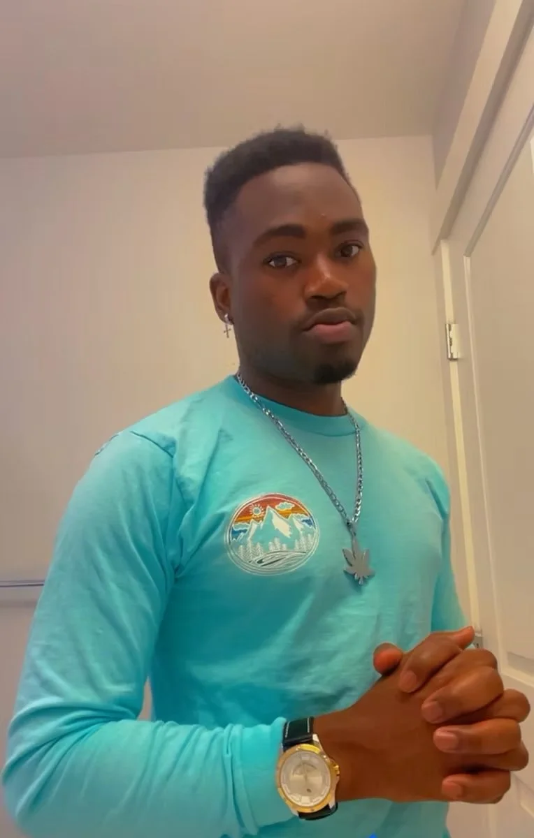 A young man with dark skin, short hair, and a serious expression, wearing a turquoise long-sleeve shirt with a mountain and sunset logo, a silver chain with a leaf-shaped pendant, a wristwatch, and an earring, standing indoors near a white door.