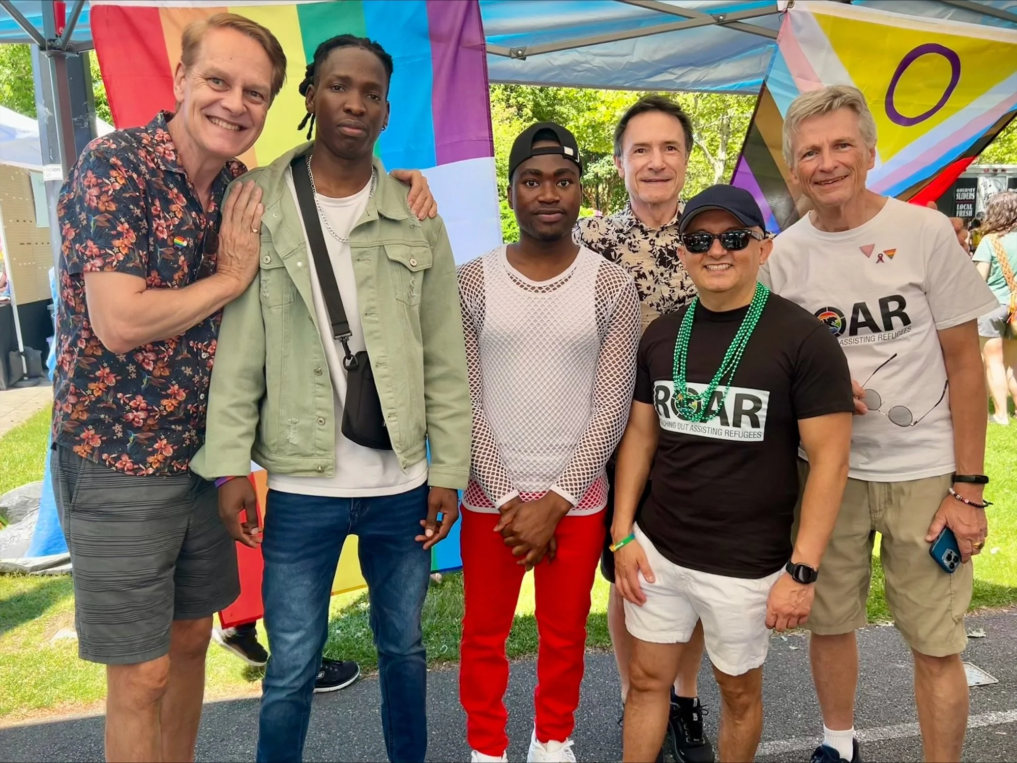 Group of six men standing together outdoors at a Pride event, with rainbow flags and colorful decorations in the background. They are smiling and dressed casually, some wearing Pride-themed clothing and accessories.