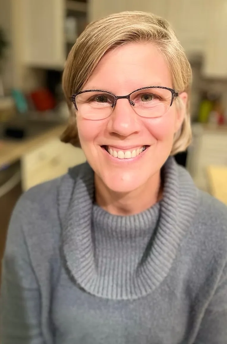 A smiling woman with short blonde hair, glasses, and a gray turtleneck sweater in a kitchen.
