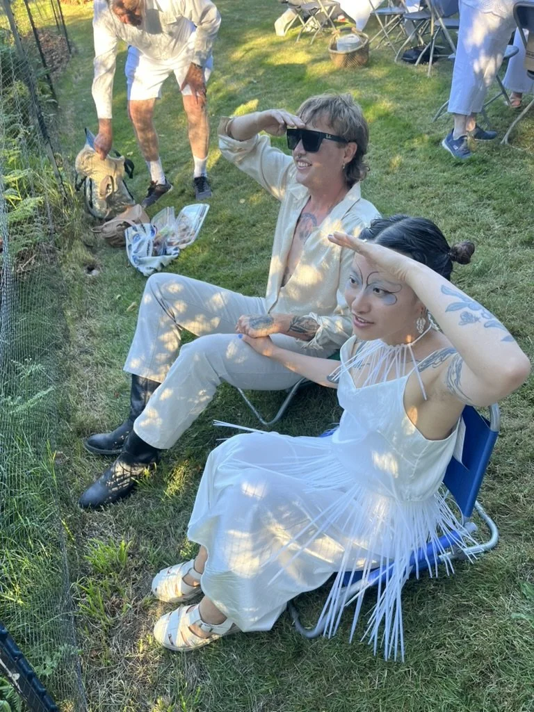 Two women sitting outdoors on chairs, dressed in white, saluting a small animal in a pen, with others standing and sitting in the background on a grassy area.