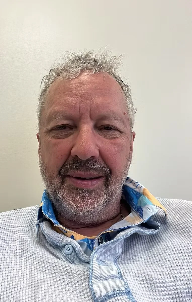 A middle-aged man with gray hair and a beard, wearing a white textured shirt with a colorful collar, sitting against a plain light-colored wall.