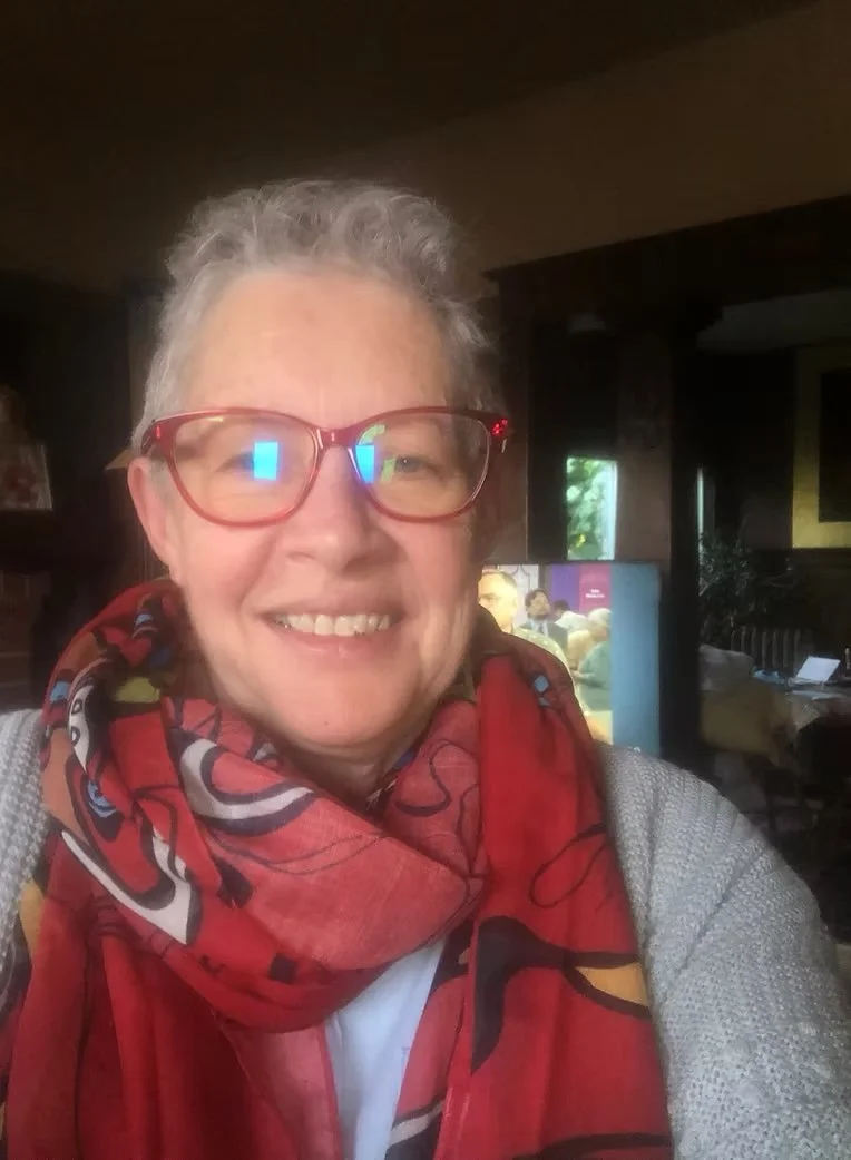 A smiling woman with short gray hair and red glasses, wearing a red and black patterned scarf and a light gray sweater, sitting indoors with a television and windows in the background.