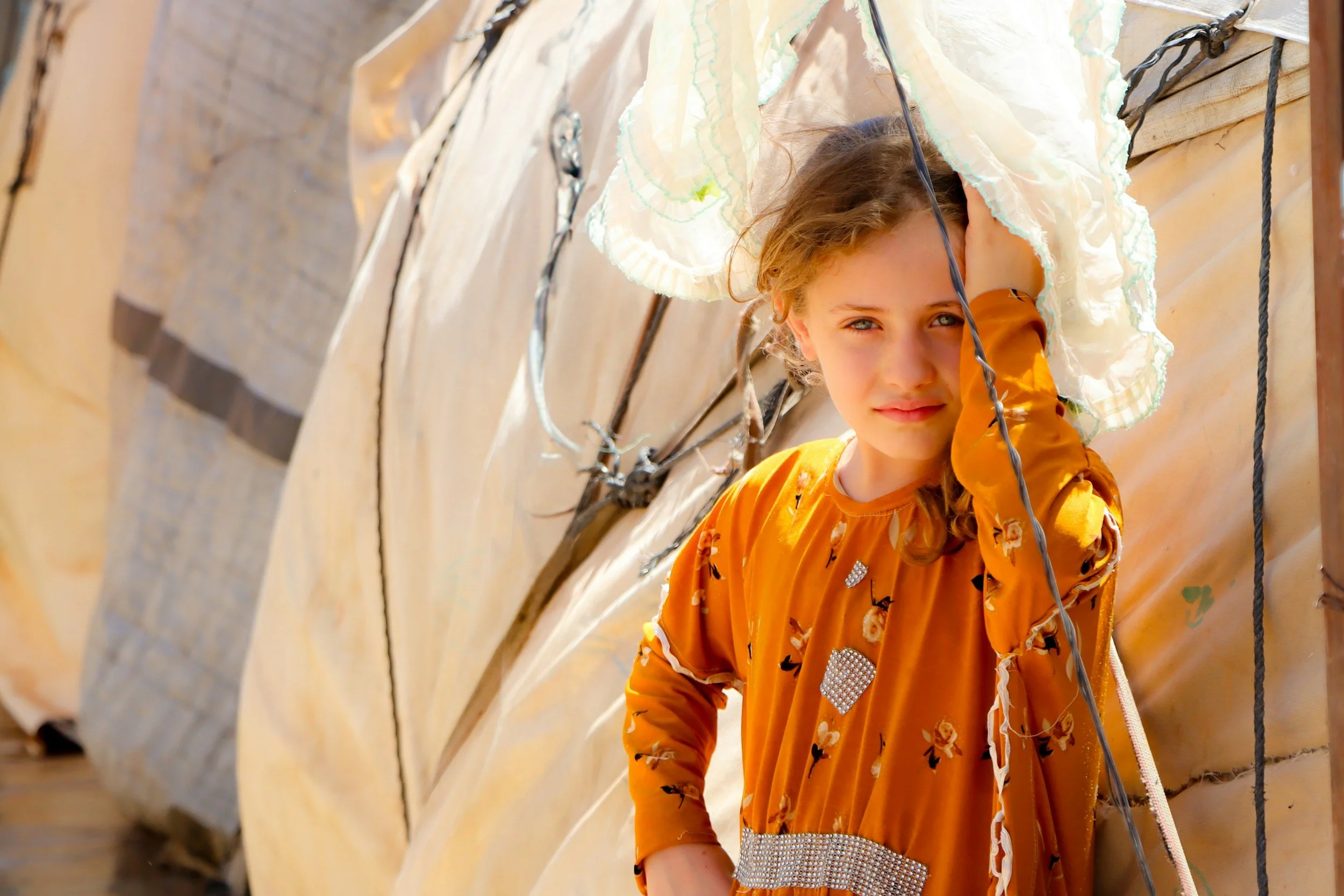 A young girl with curly hair and a yellow dress standing beside a large tarp or tent, leaning her head on her hand, with a calm expression.