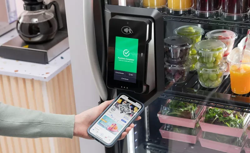 Person making a contactless payment with a smartphone at a vending machine containing pre-packaged salads, fruit cups, and beverages.