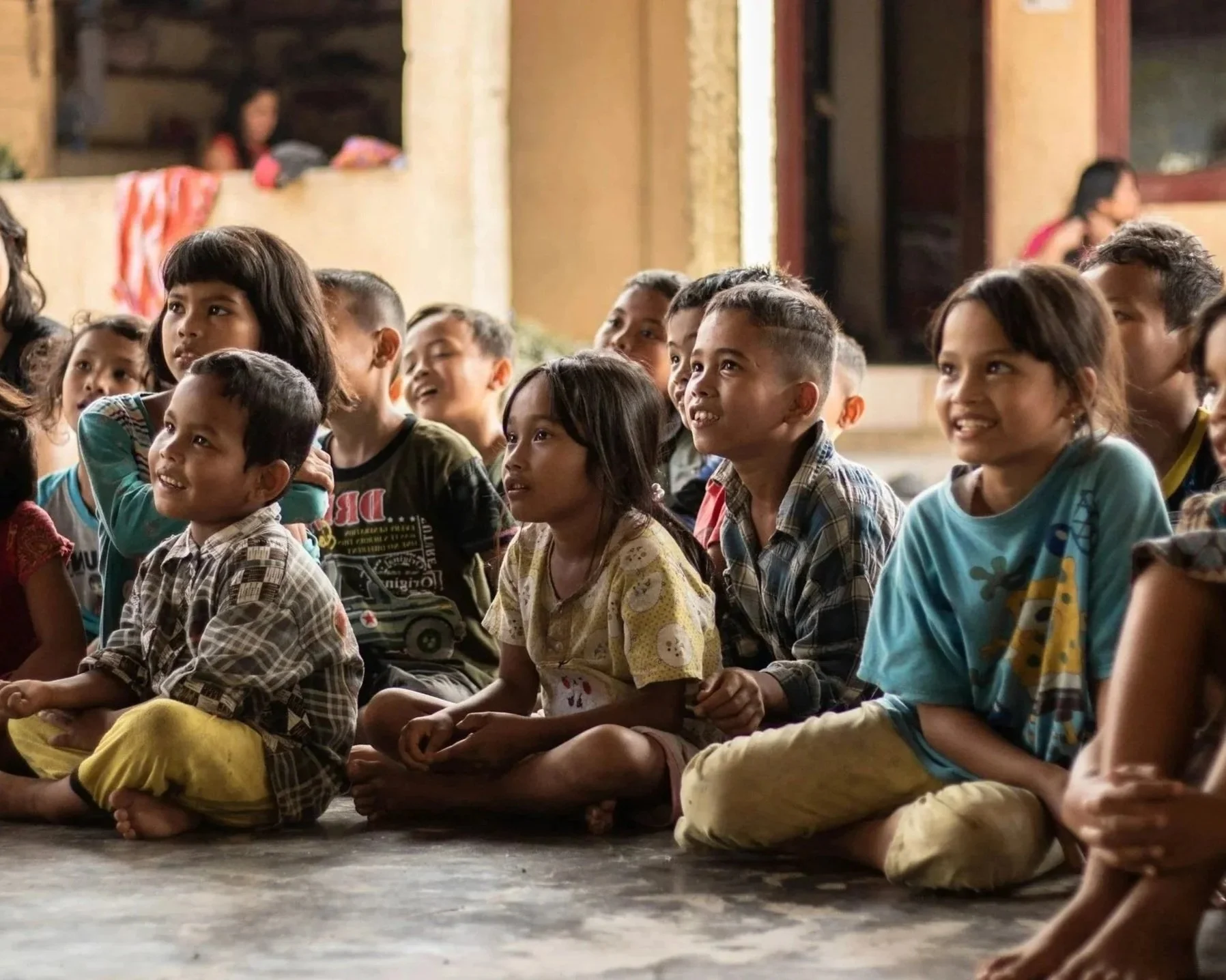 A group of children sitting on the floor, watching something with smiles and interest, inside a rustic room with beige walls and windows.