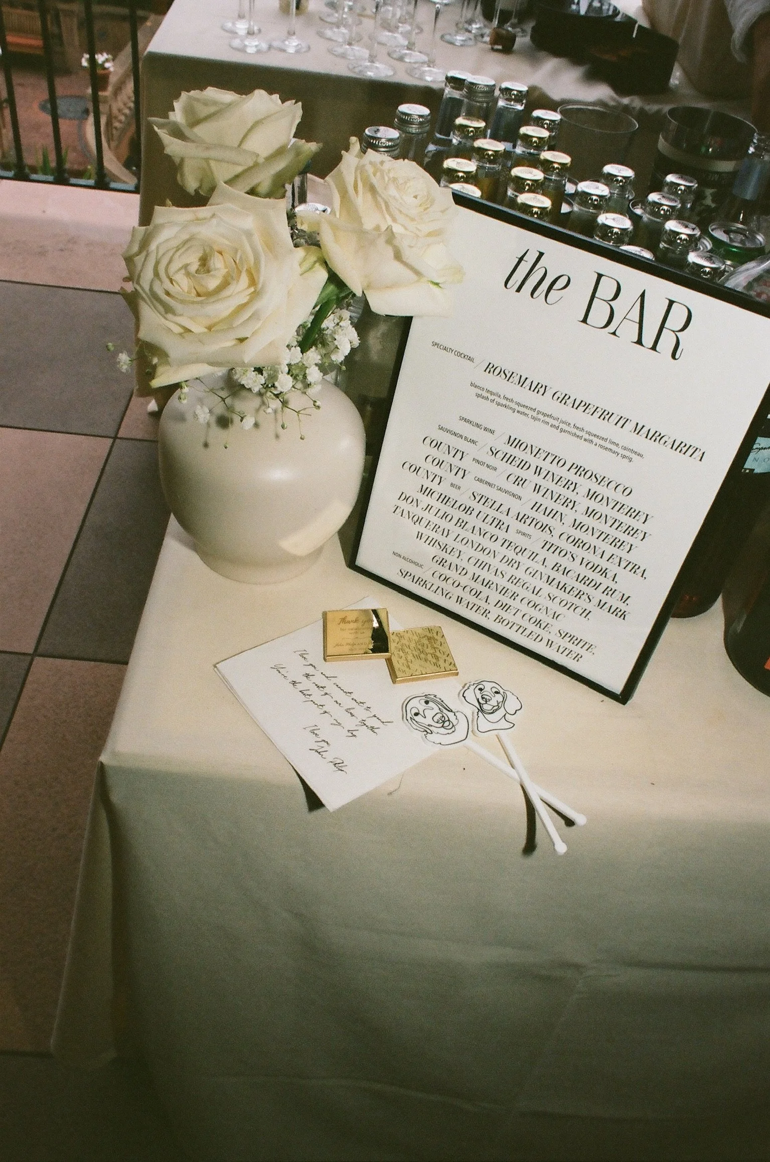 A table with a vase of white roses, a menu board for a bar, and some small decorative drawings. The menu lists various cocktails and beverages.