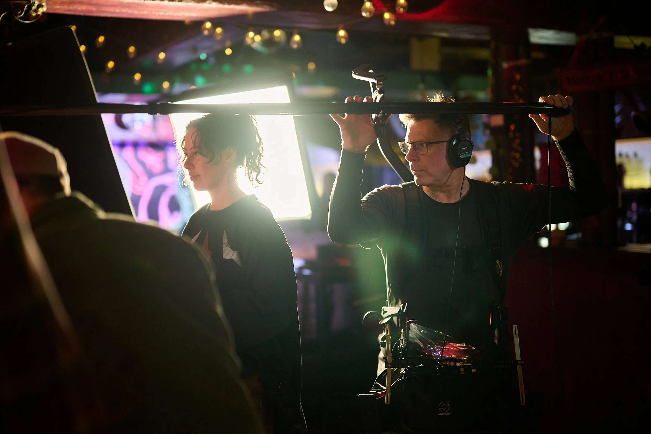 Sound recordist Chris Ward recording sound beside an extra while filming the Statistics New Zealand commercial 'Data Helps Us Do in a dimly lit bar