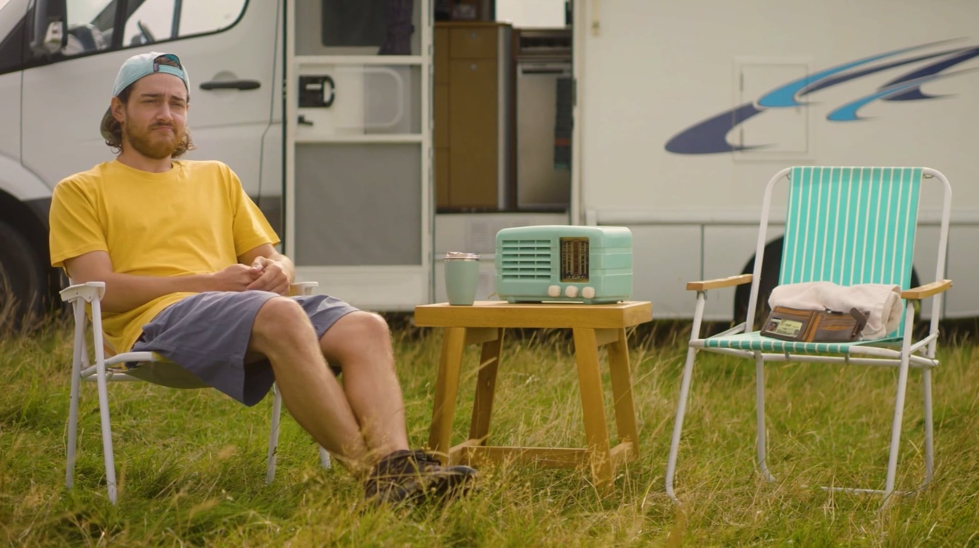 Waka Kotahi advertisement for renewing licenses. Man sitting outdoors on a folding chair at a campsite, wearing a yellow shirt, gray shorts, and a backwards cap, with a vintage radio and a cup on a small wooden table, and an RV in the background.