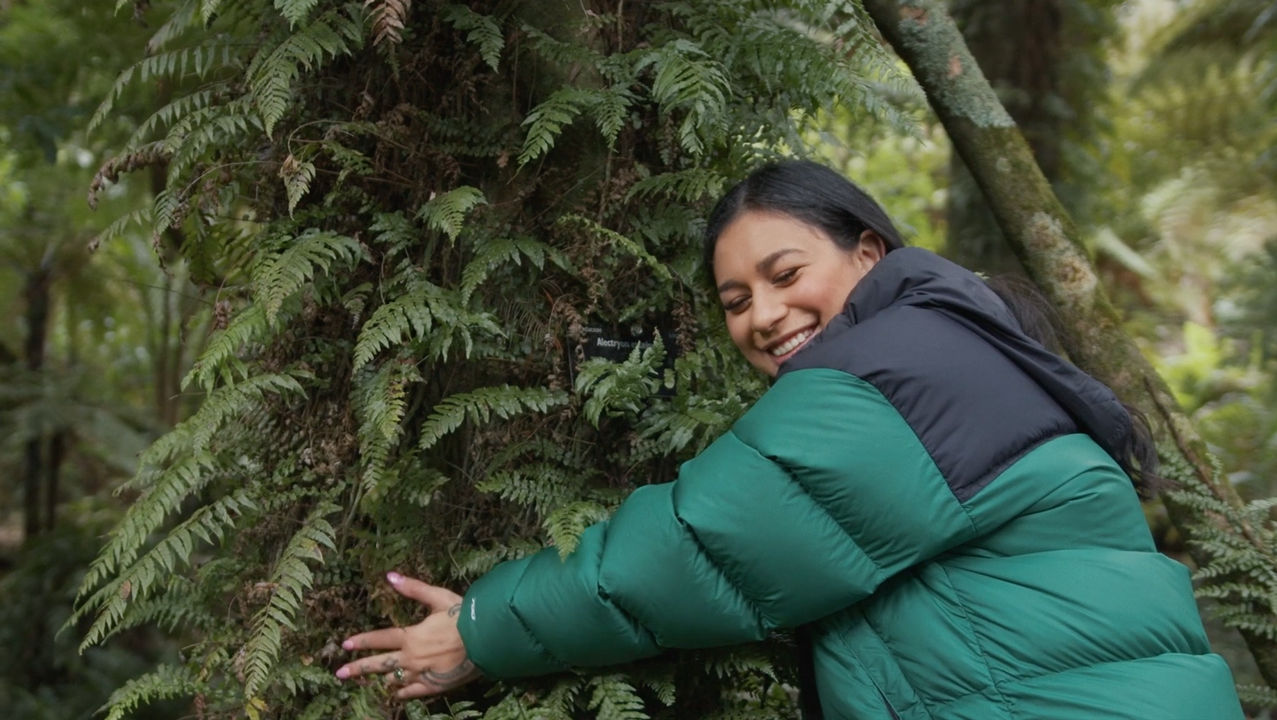 Green Party of Aotearoa New Zealand MP Tamatha Paul documentary - smiling embracing a fern-covered tree trunk in a forest, wearing a teal and black puffy jacket.