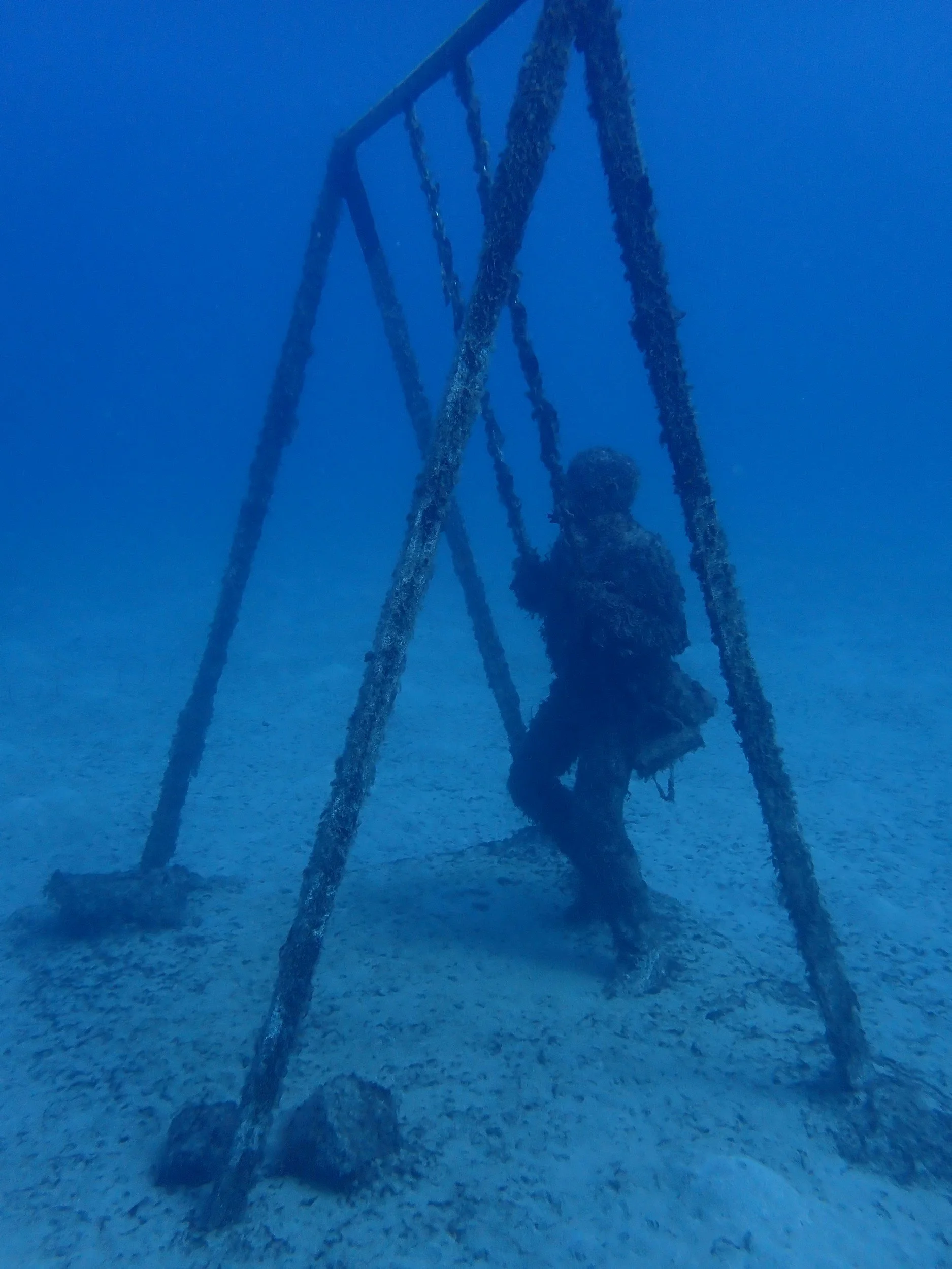Silhouette di una persona su una sedie a sdraio sotto l'acqua, visibile in profondità marina con fondo sabbioso.