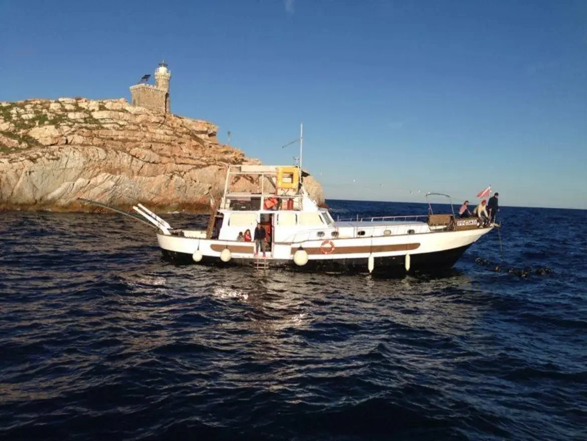 Una barca a vela bianca in mare vicino a una scogliera con un faro in cima, con alcune persone a bordo.