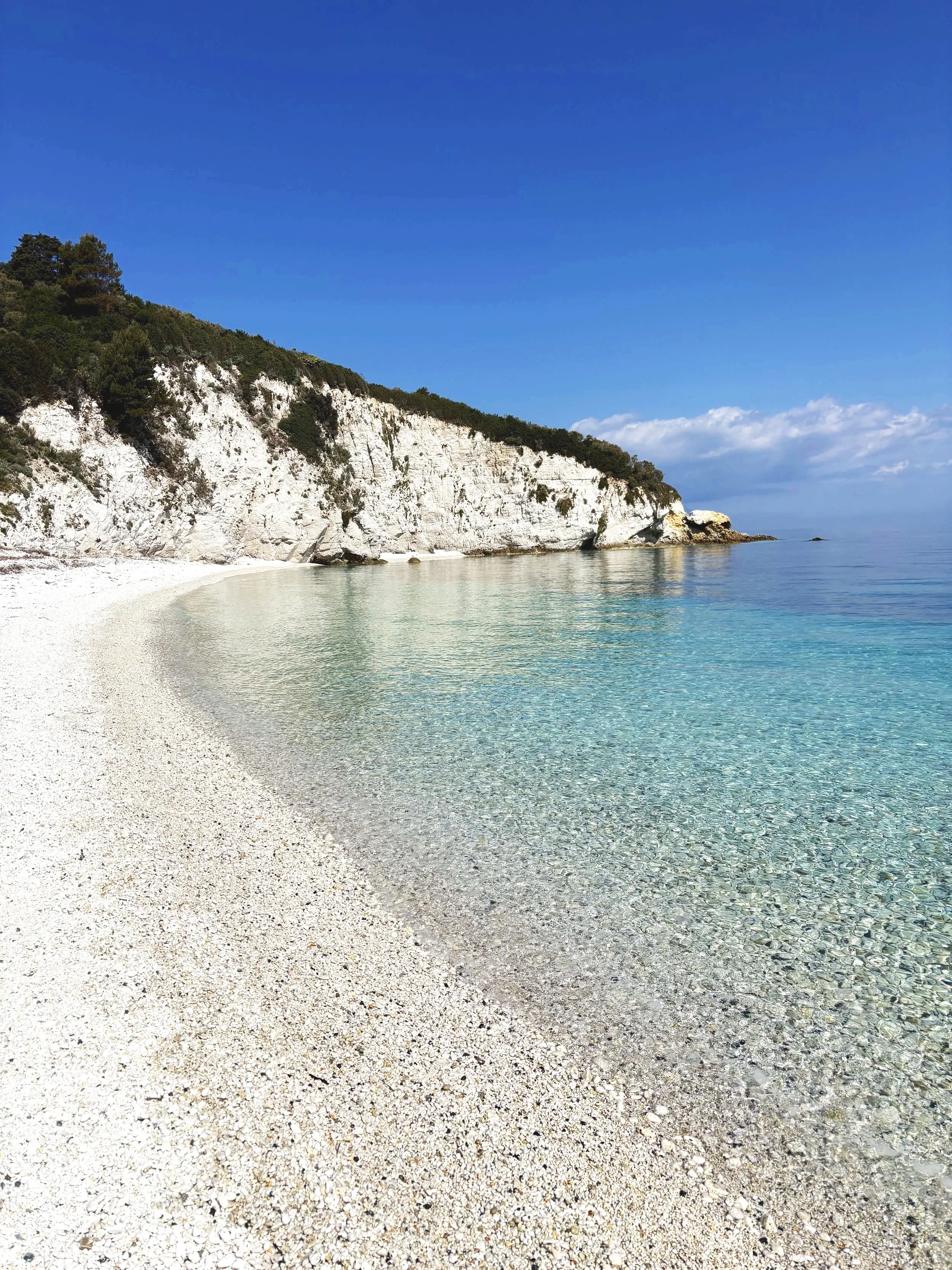 Spiaggia di ghiaia con acque cristalline, scogli bianchi e una scogliera ricoperta di vegetazione verde sotto un cielo azzurro con alcune nuvole.
