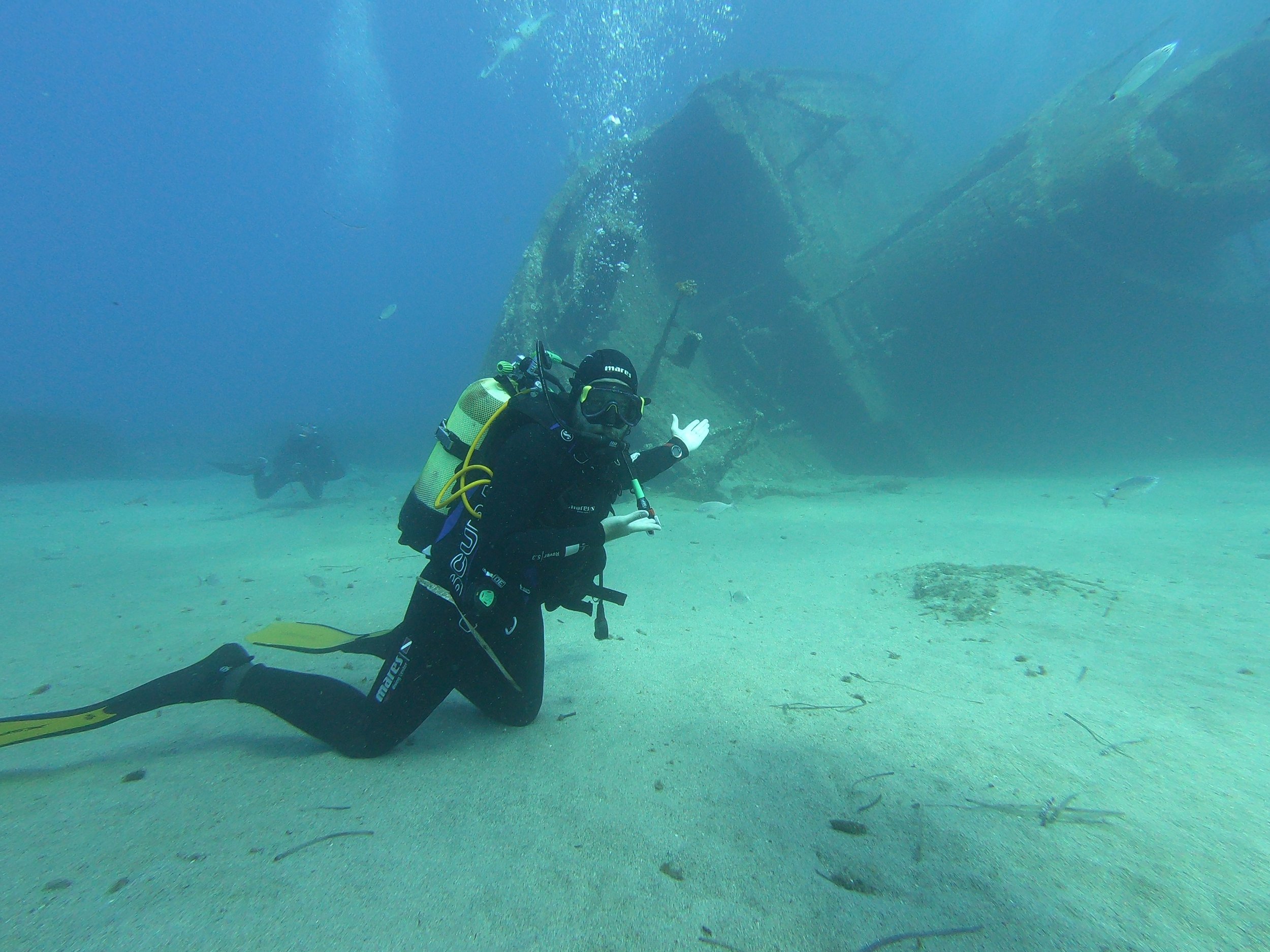 Subacqueo con attrezzatura completa, in ginocchio sul fondale sabbioso, osservando un relitto sommerso, con alcuni pesci e una nave affondata visibile sullo sfondo.