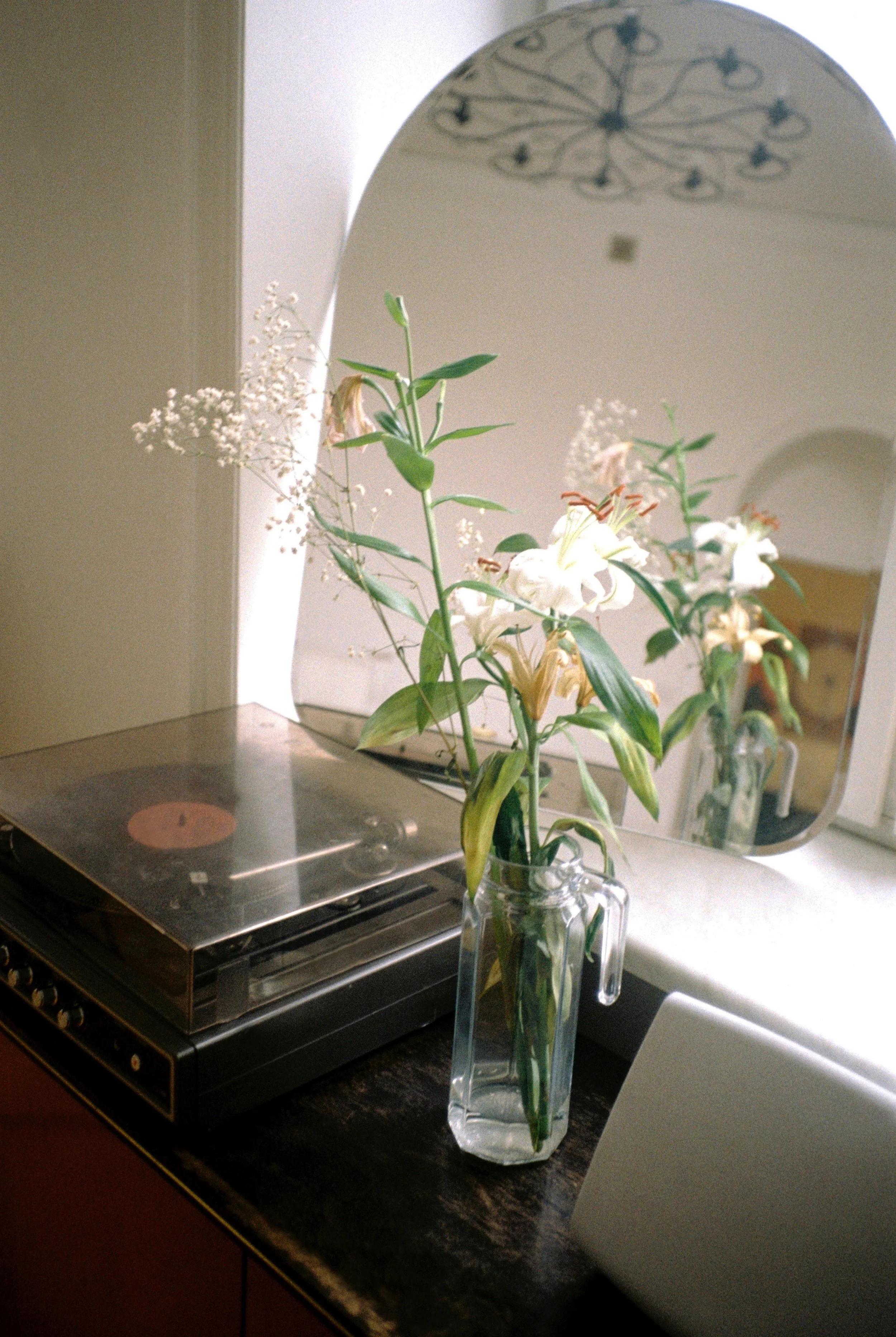 Vase with white lilies and greenery on a black countertop next to a turntable and a mirror reflecting the flowers in a room.