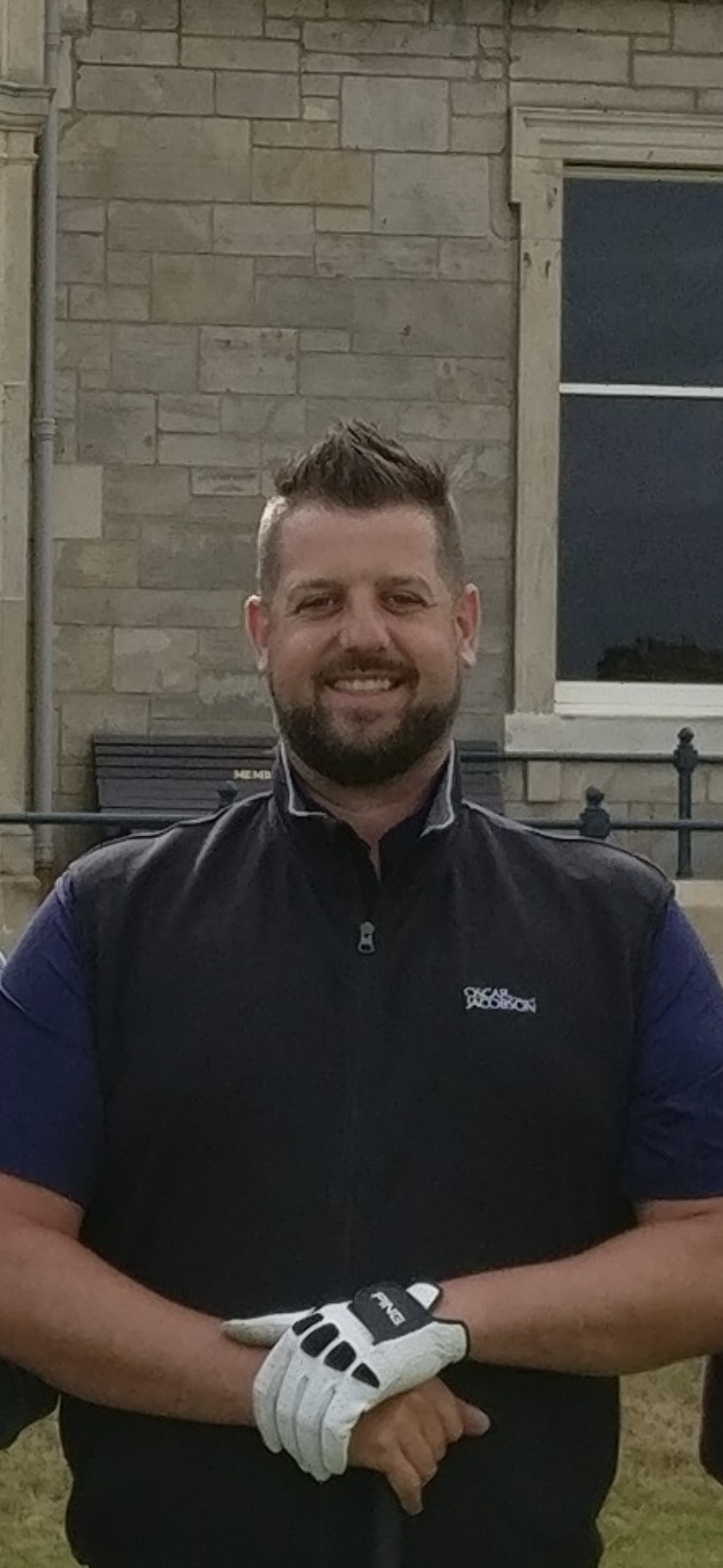 A smiling man with dark hair and a beard wearing a black jacket over a blue shirt, standing outdoors in front of a brick wall and window, with his hands crossed in front of him, one hand wearing a white golf glove.