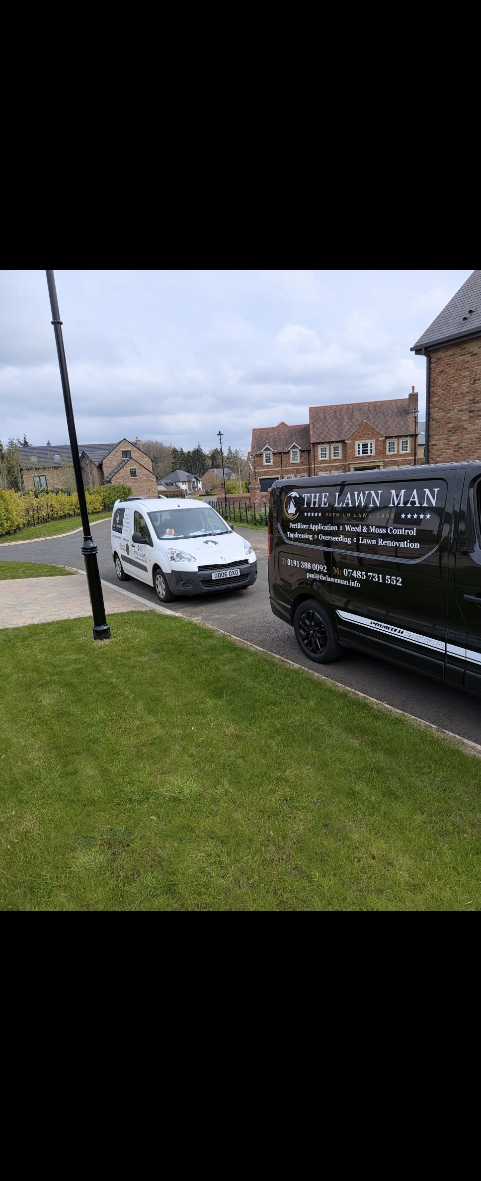 A residential driveway with two parked work vehicles from The Lawn Man. One white van and one black van, both with company branding. Surrounding houses are brick with tiled roofs, with a grassy area and a streetlamp nearby.
