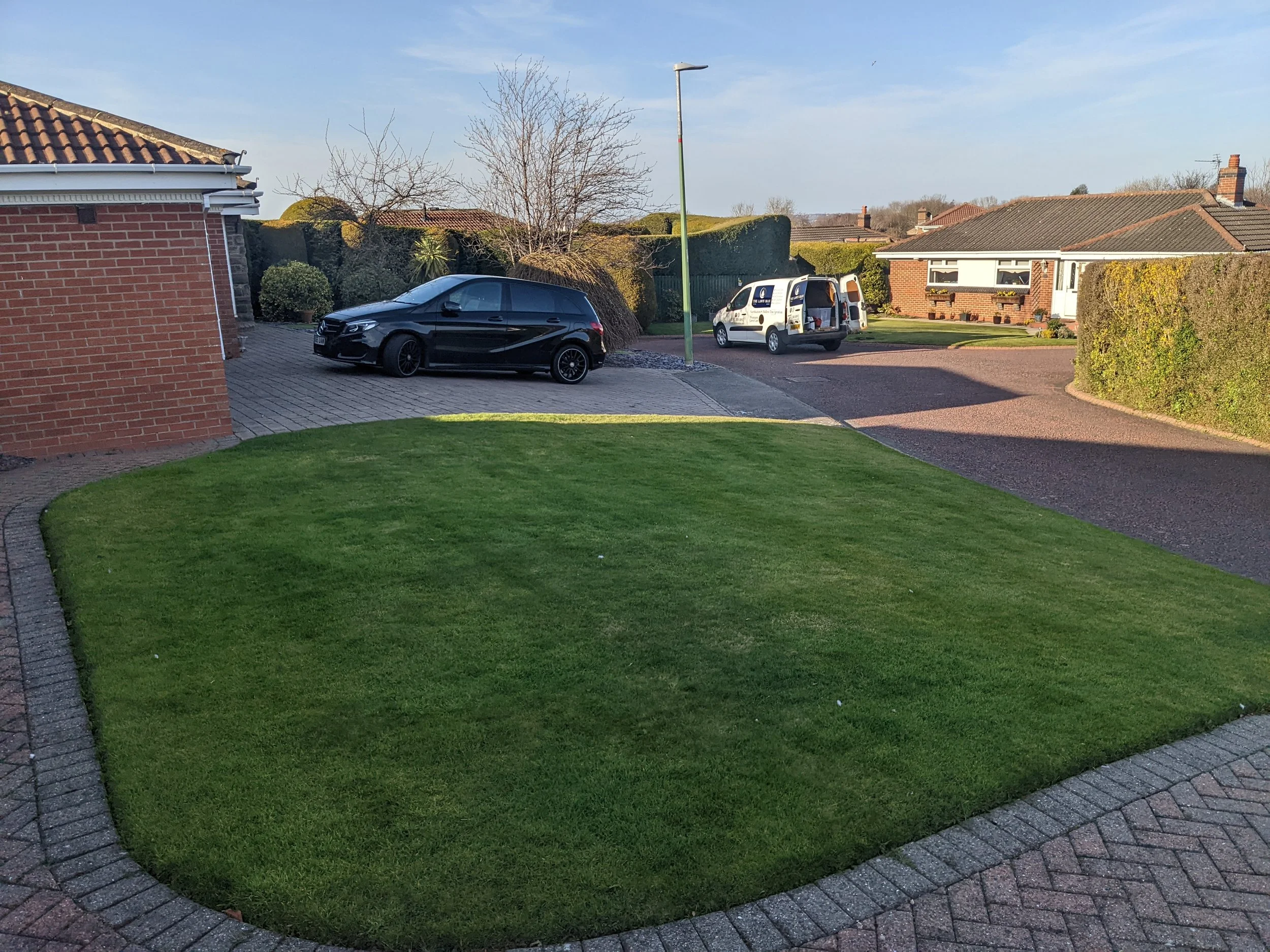 A suburban house driveway with a black Mercedes-Benz parked on a cobblestone path, a white work van with open doors, and neighboring houses with lawns and hedges in the background on a sunny day.