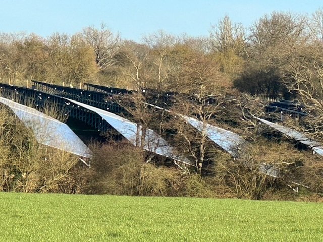 A bridge over a landscape, with solar panels installed on its sides, surrounded by trees and a grassy foreground.