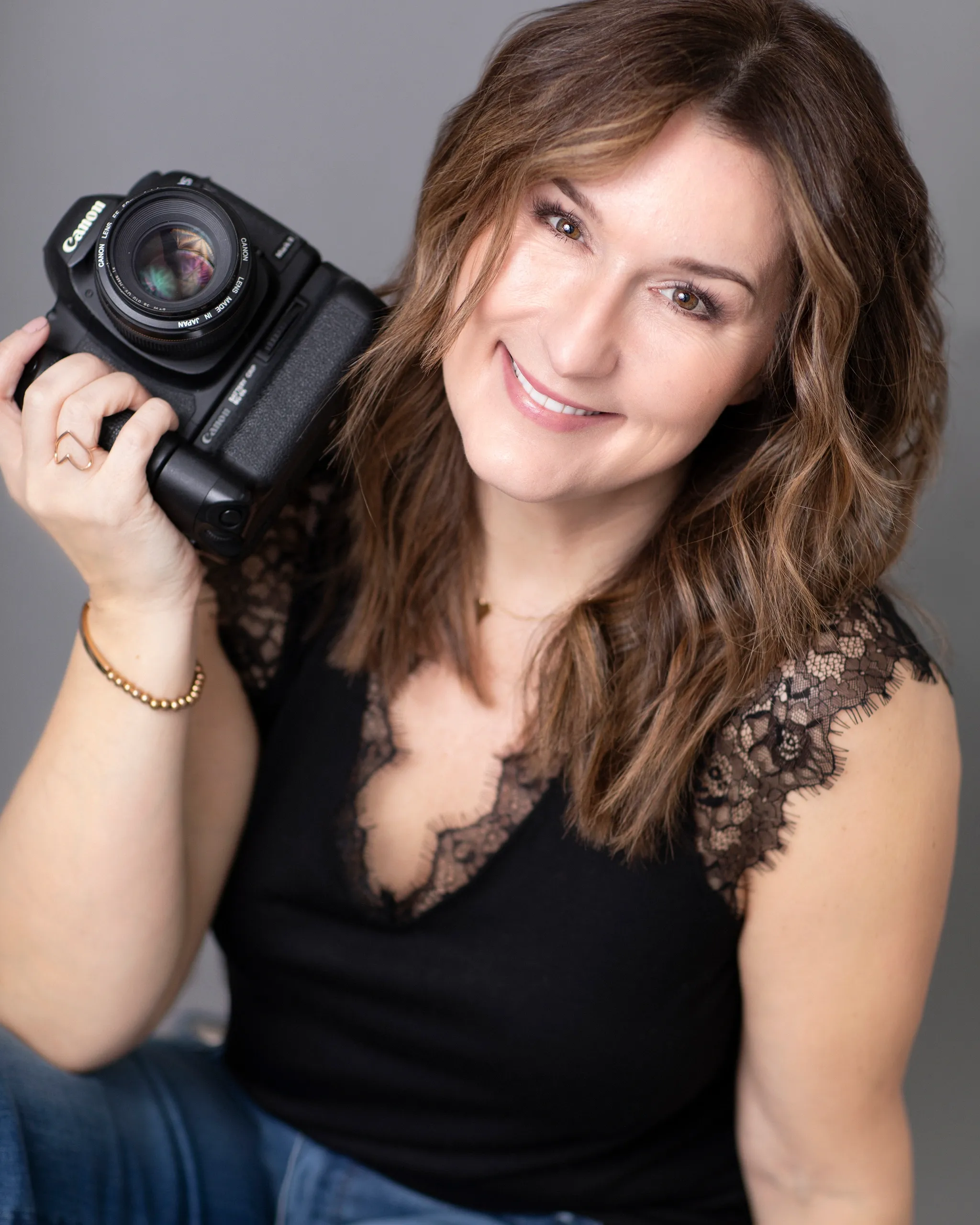 A woman with brown, wavy hair, smiling, holding a camera near her face.