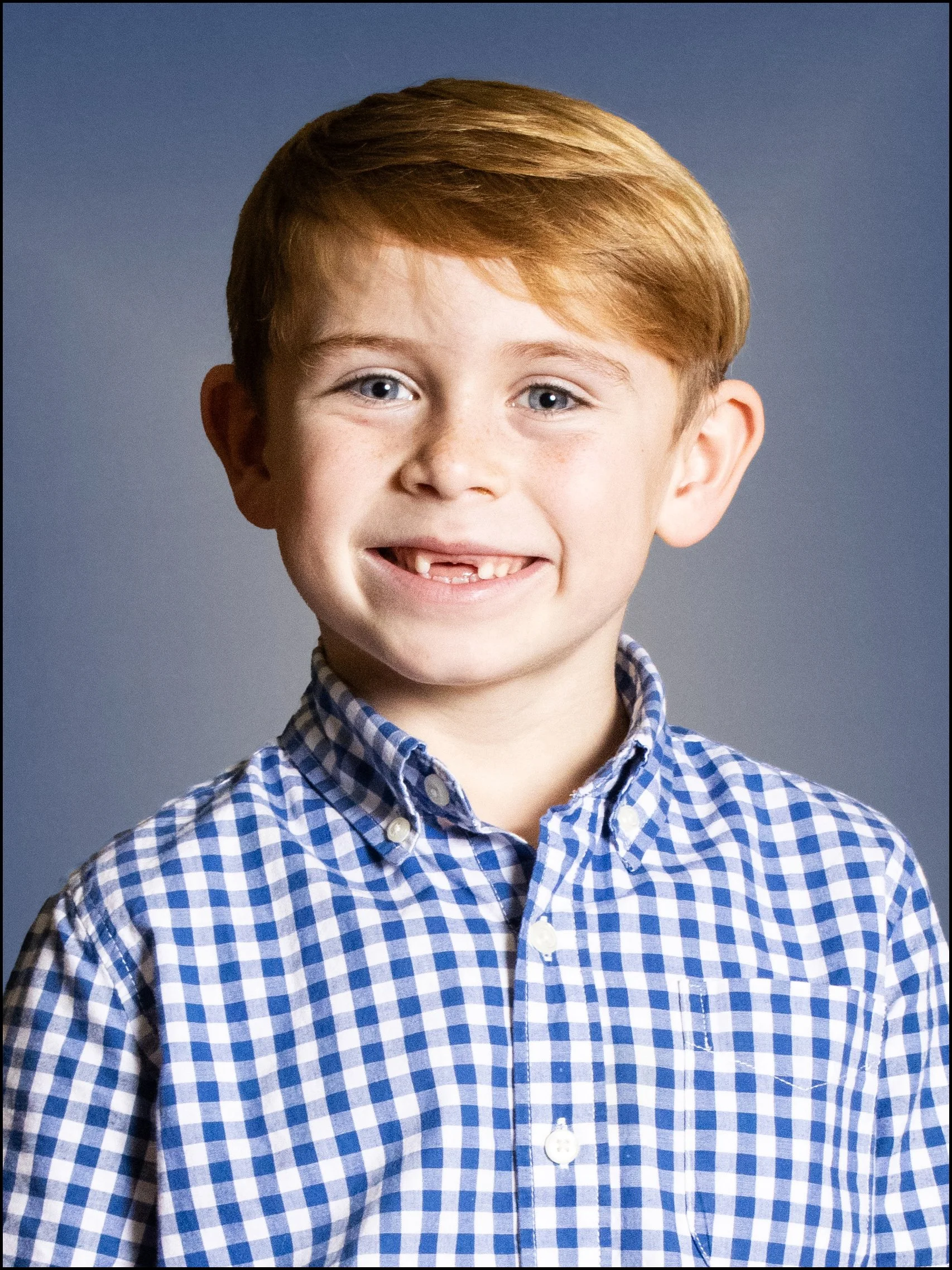 A young boy with red hair, blue eyes, and freckles smiling, wearing a blue and white checked shirt, against a gray background.
