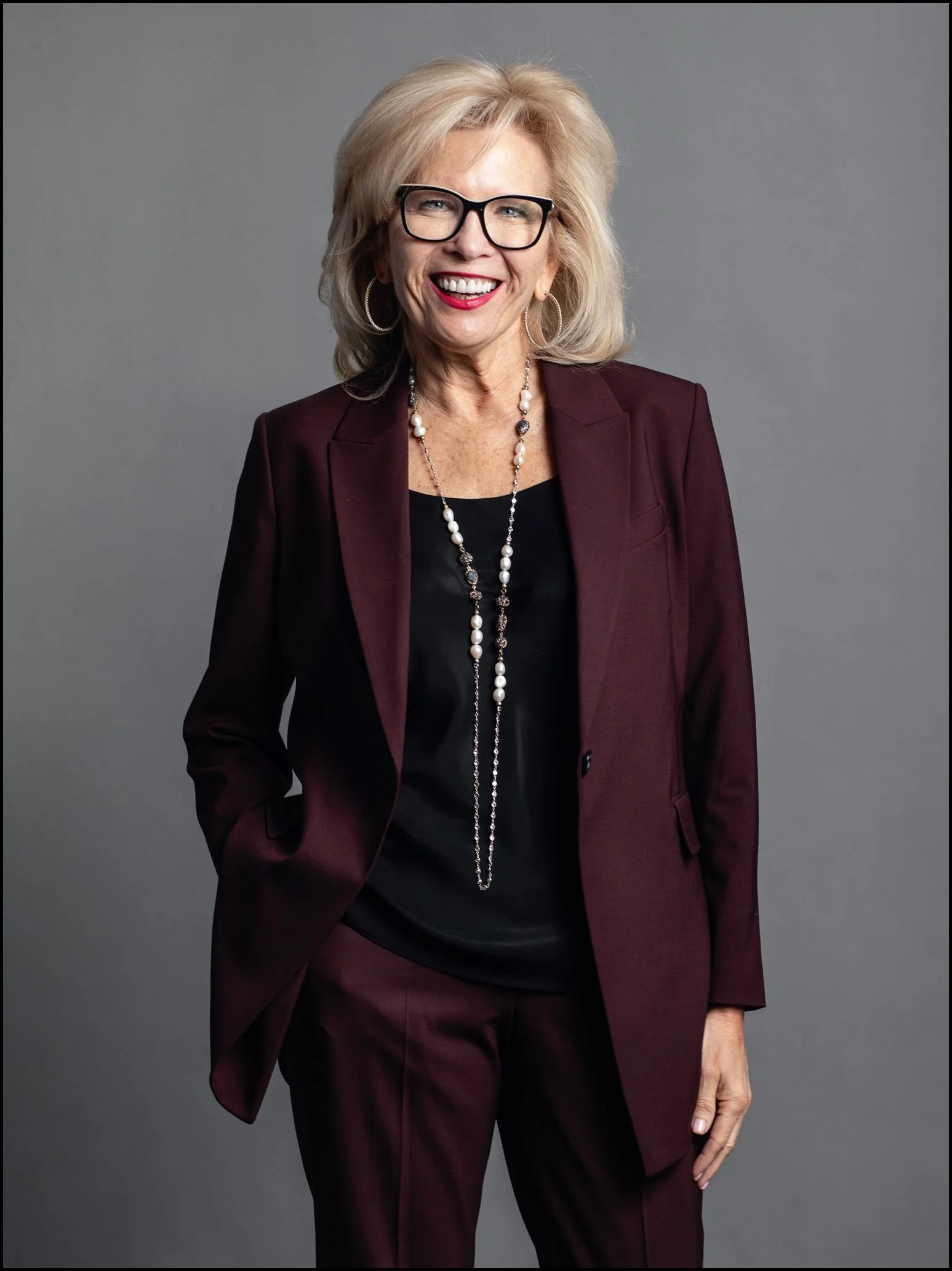 A cheerful elderly woman with blonde hair, wearing glasses, a maroon suit with matching pants, a black top, and accessorized with hoop earrings and layered pearl necklaces, posing against a plain gray background.