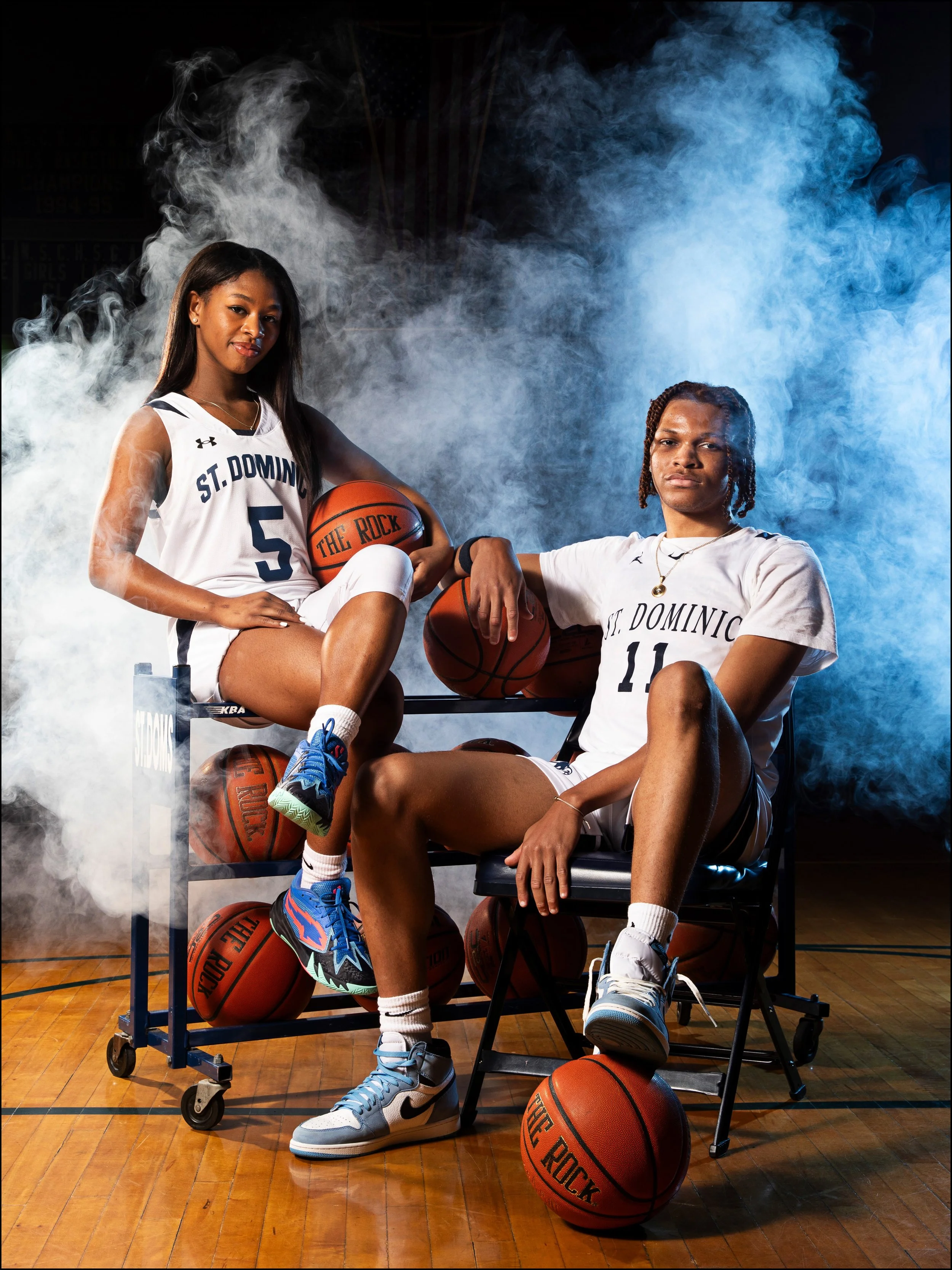 Two female basketball players in white jerseys with 'St. Dominic' and numbers 5 and 11, posing with basketballs on a gymnasium wooden floor, surrounded by smoke effects.
