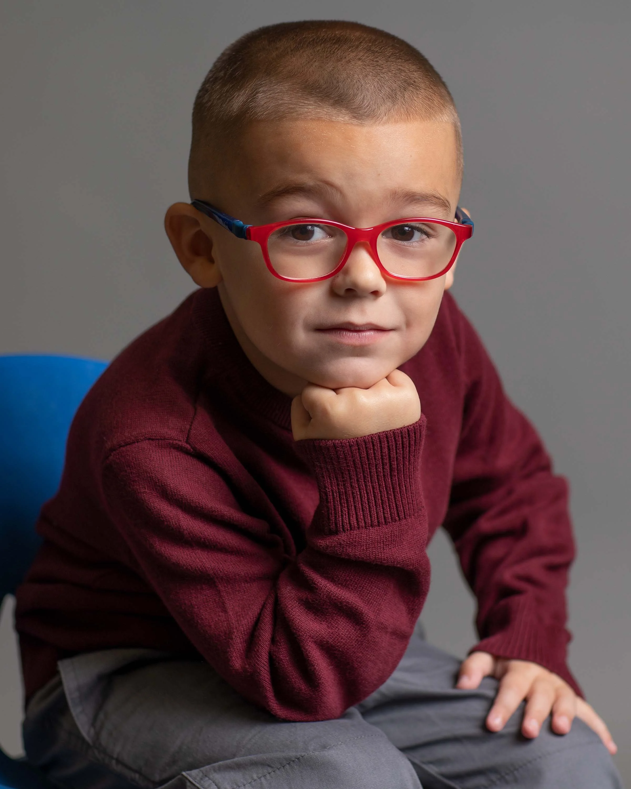 A young boy with short hair wearing red glasses, sitting on a blue chair, resting his chin on his hand, wearing a maroon sweater.