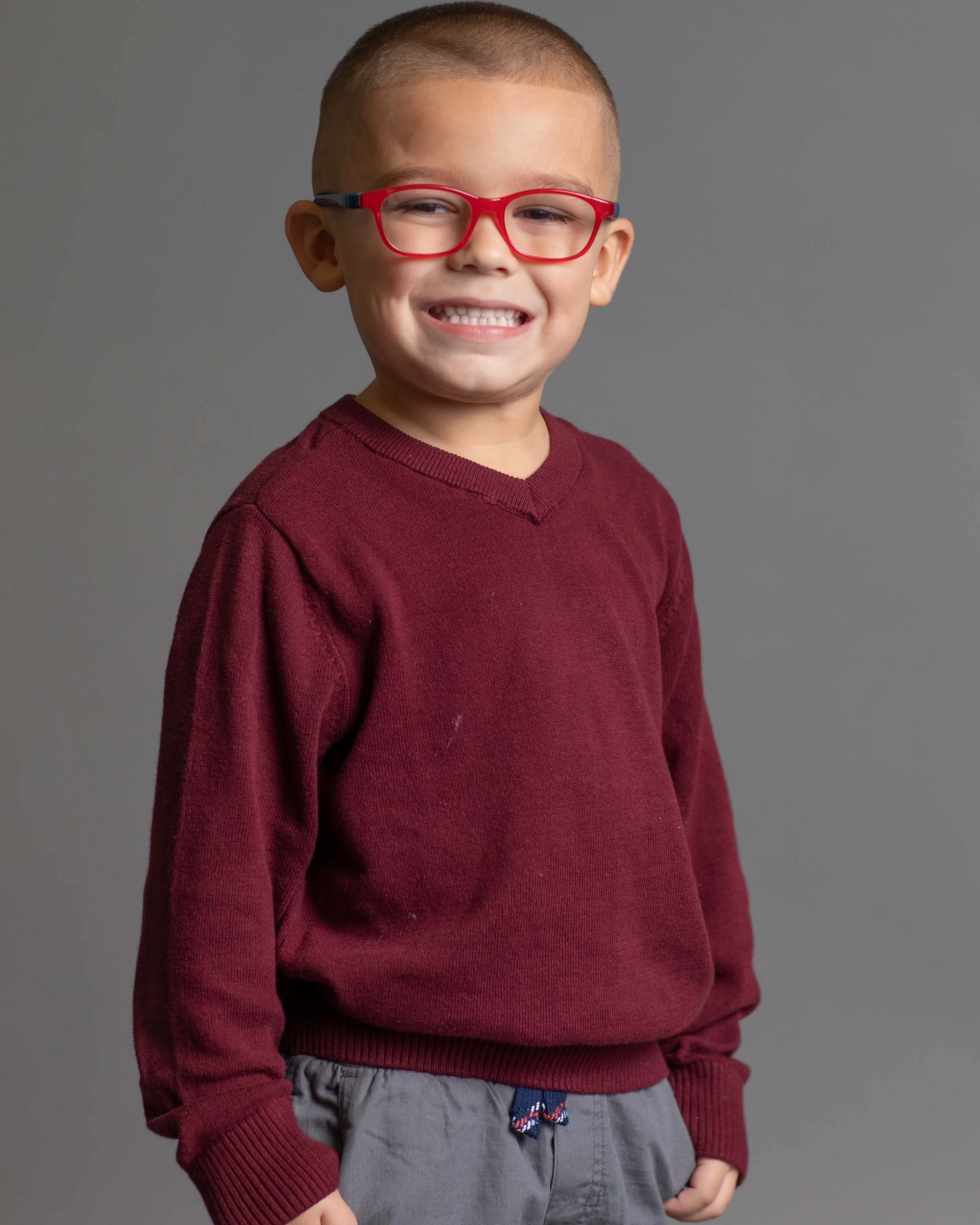 A young boy with short hair, red glasses, and a big smile, wearing a maroon sweater and gray pants, standing against a plain gray background.
