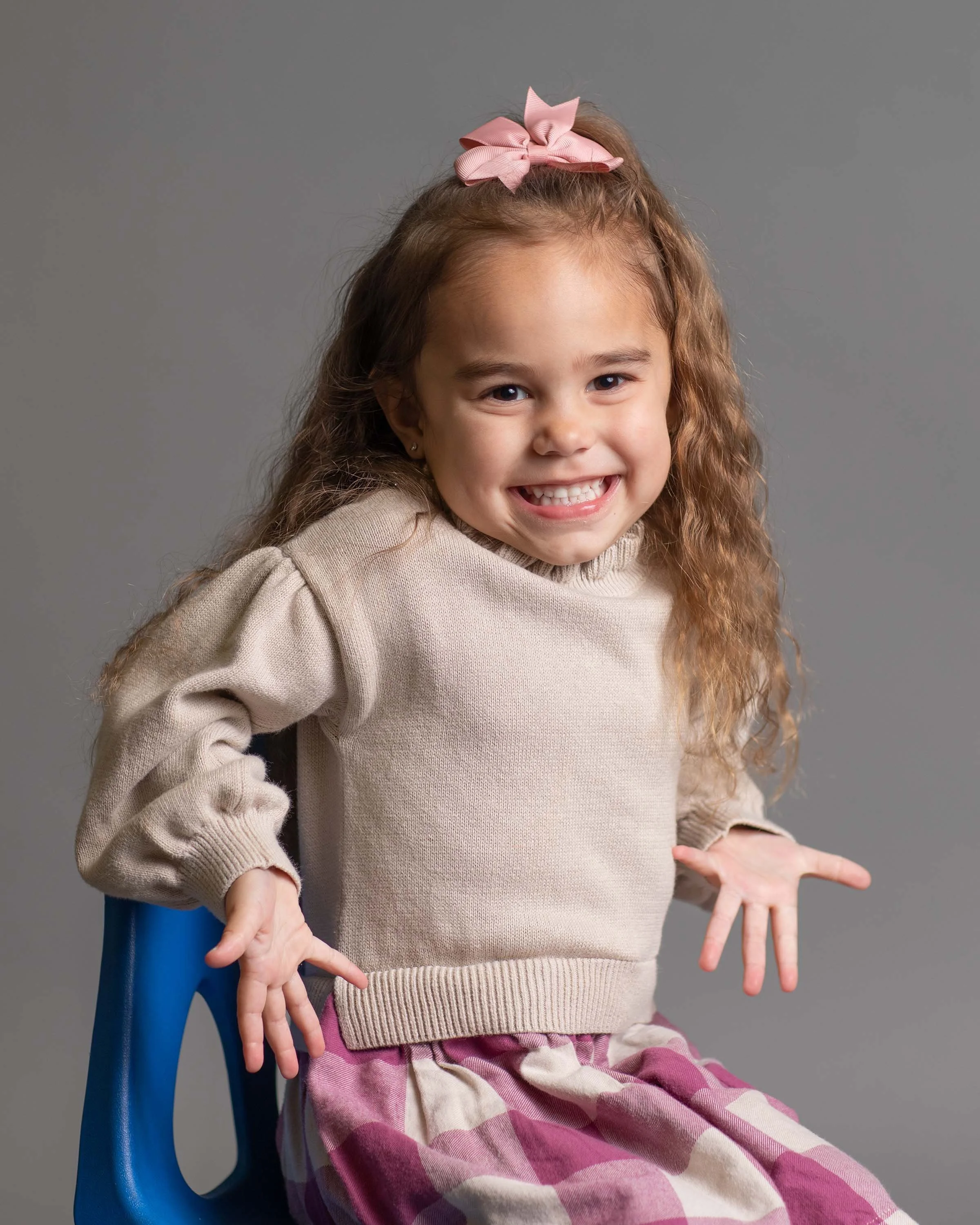 A young girl with curly hair seated on a blue chair, smiling broadly, wearing a beige sweater, pink and purple plaid skirt, with a pink bow in her hair, against a gray background.