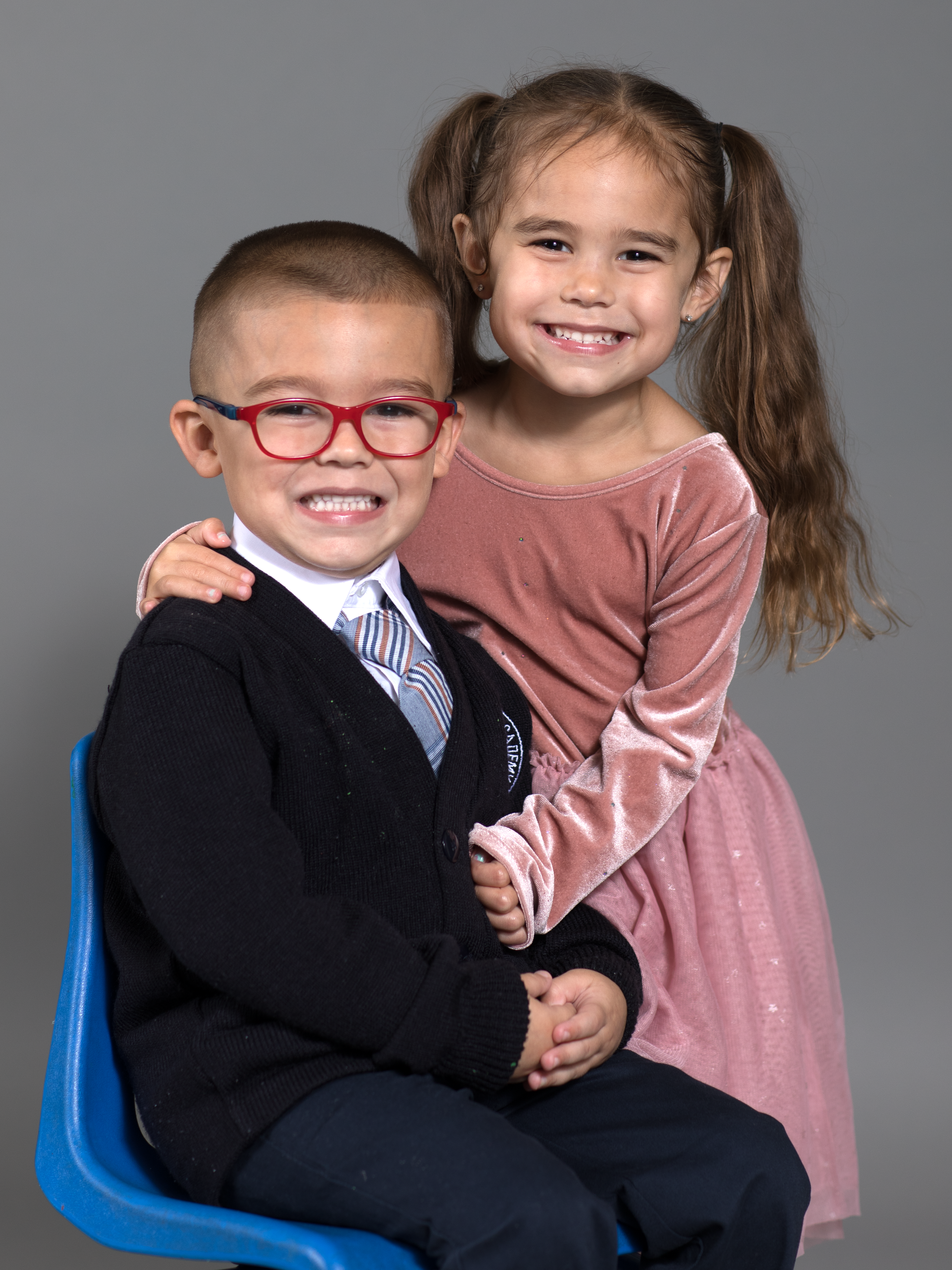 A young boy wearing glasses, a white shirt, a vest, and a tie is sitting on a blue chair, with a young girl in a pink dress standing behind him, both smiling at the camera against a plain gray background.