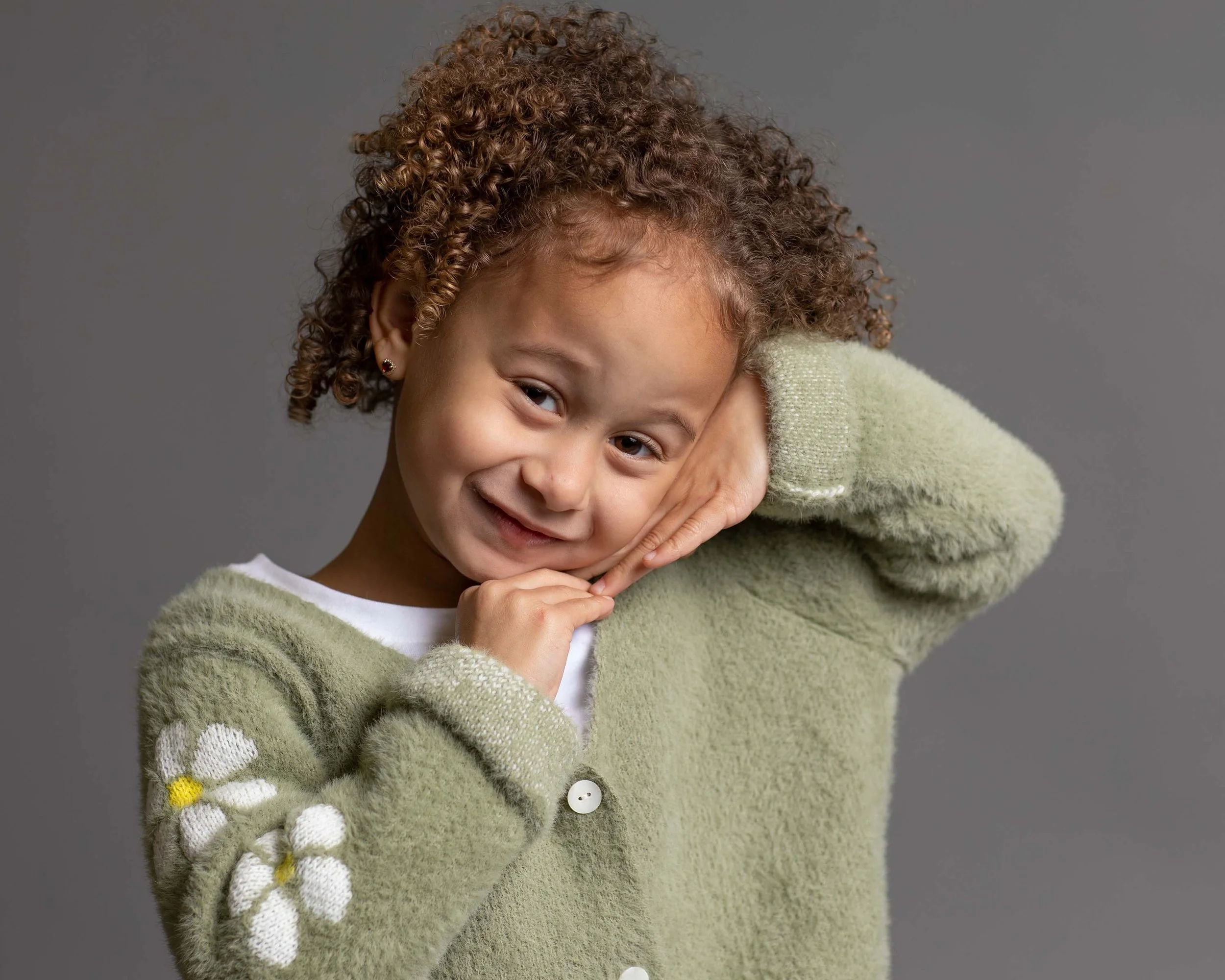 A young girl with curly brown hair, smiling and resting her head on her hand, wearing a green sweater with daisy embroidery on the sleeve against a gray background.