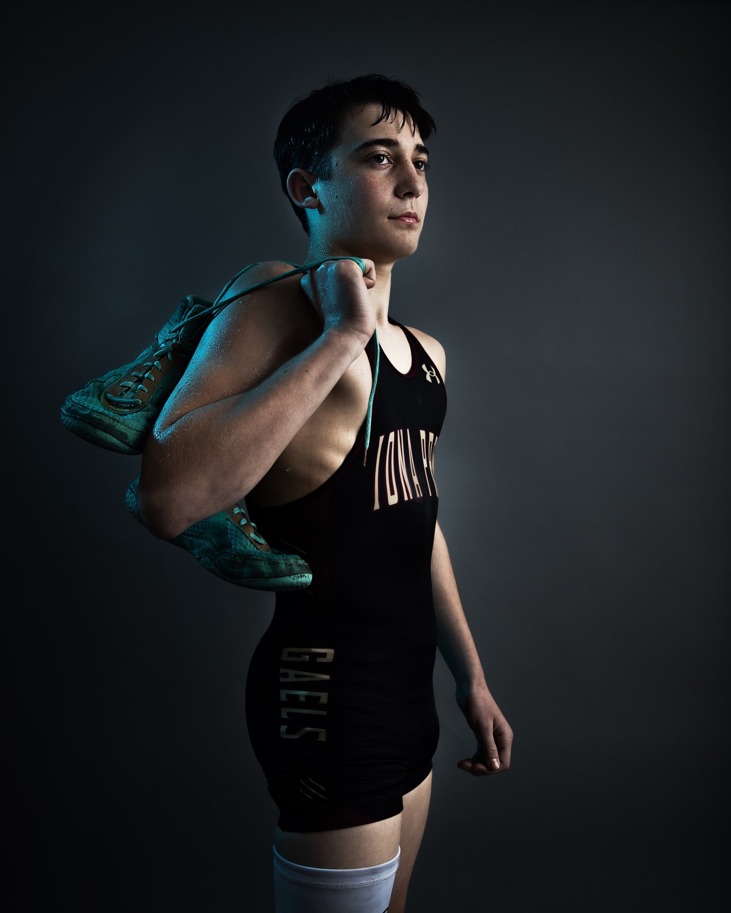 Young female athlete in athletic gear with a shoe over her shoulder, standing against a dark background.