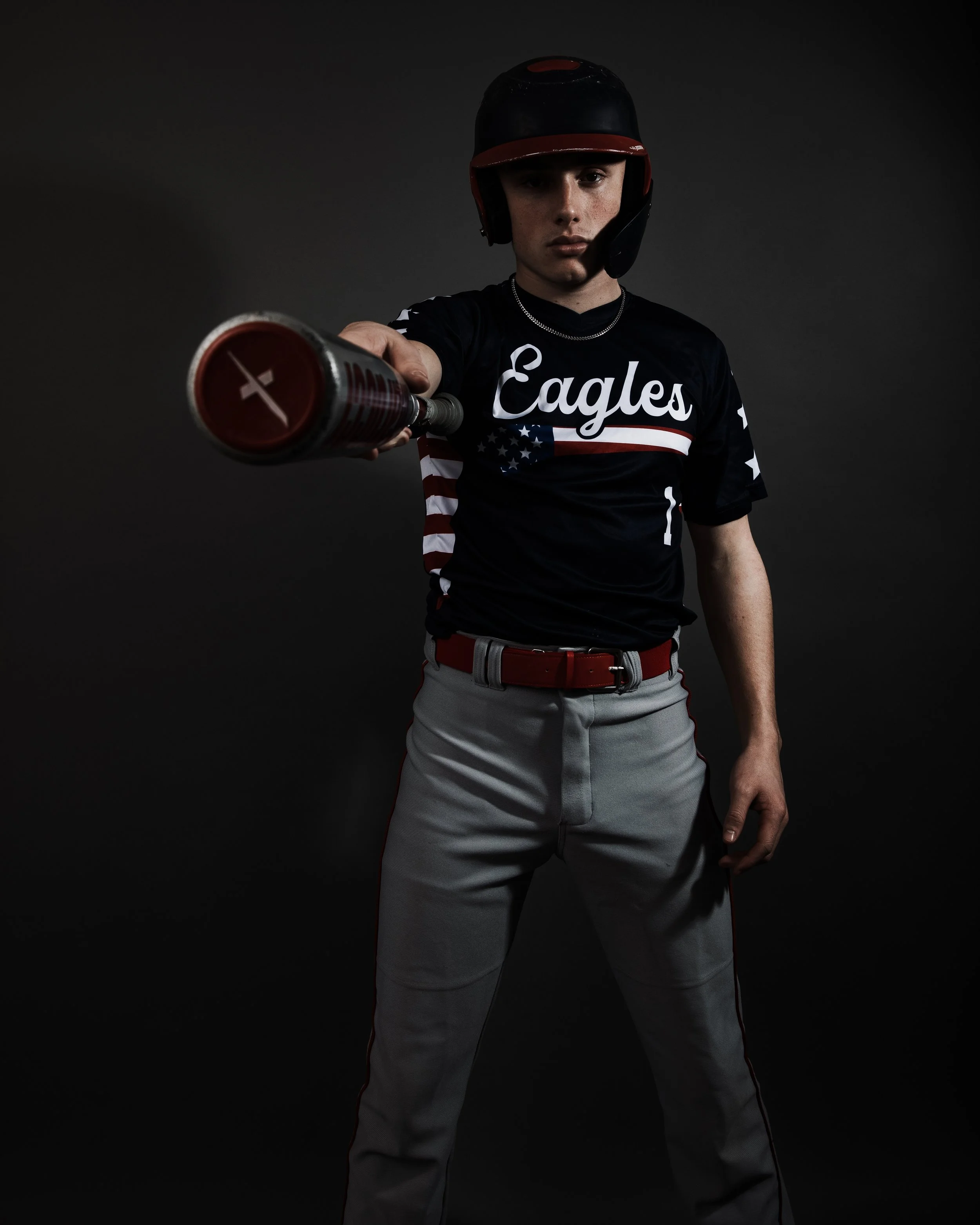 A young baseball player in uniform, wearing a helmet, holding a bat pointed toward the camera, against a dark background.