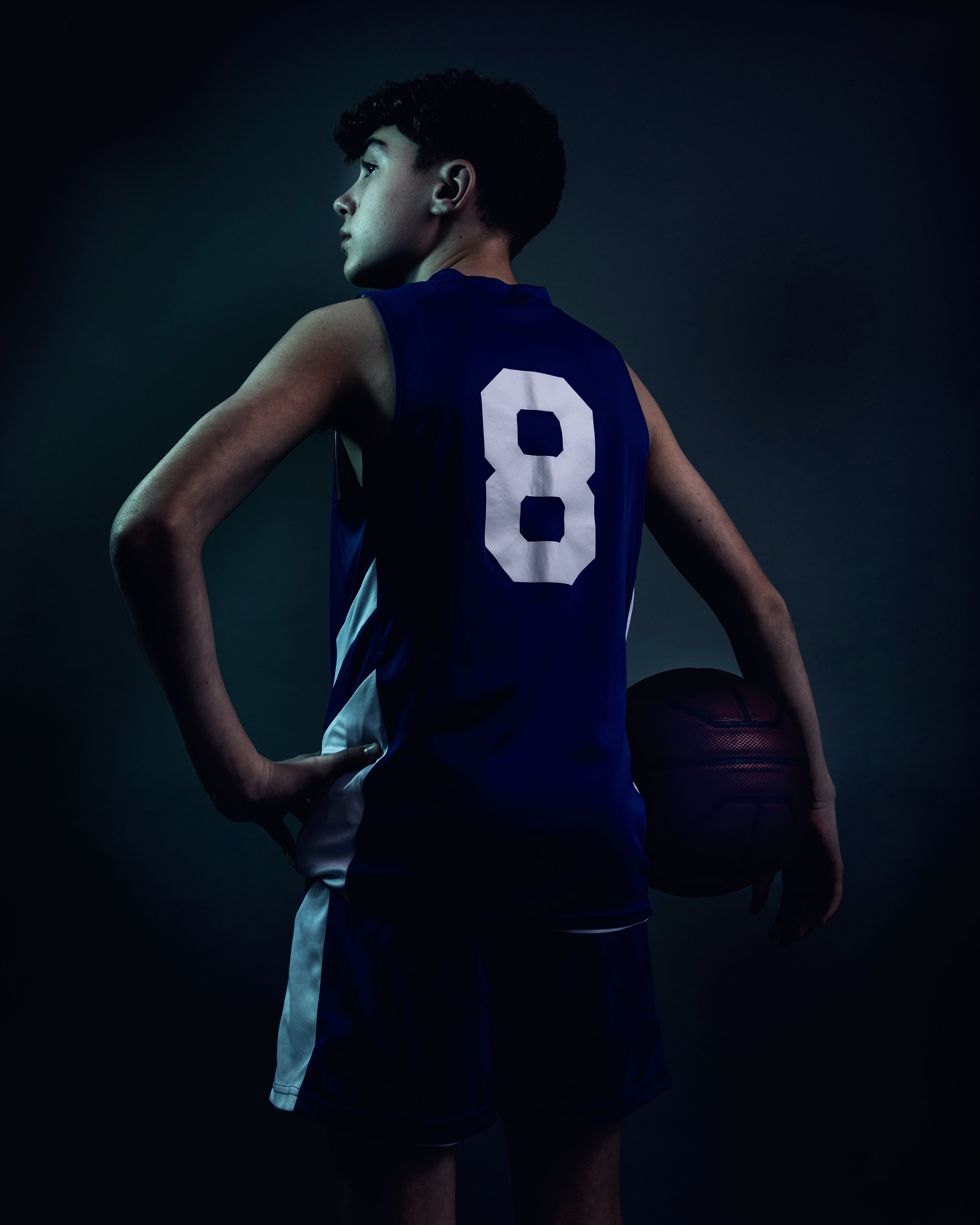 A young male basketball player with curly dark hair, wearing a blue jersey with the number 8, holding a basketball behind his right thigh, standing in profile against a dark background.