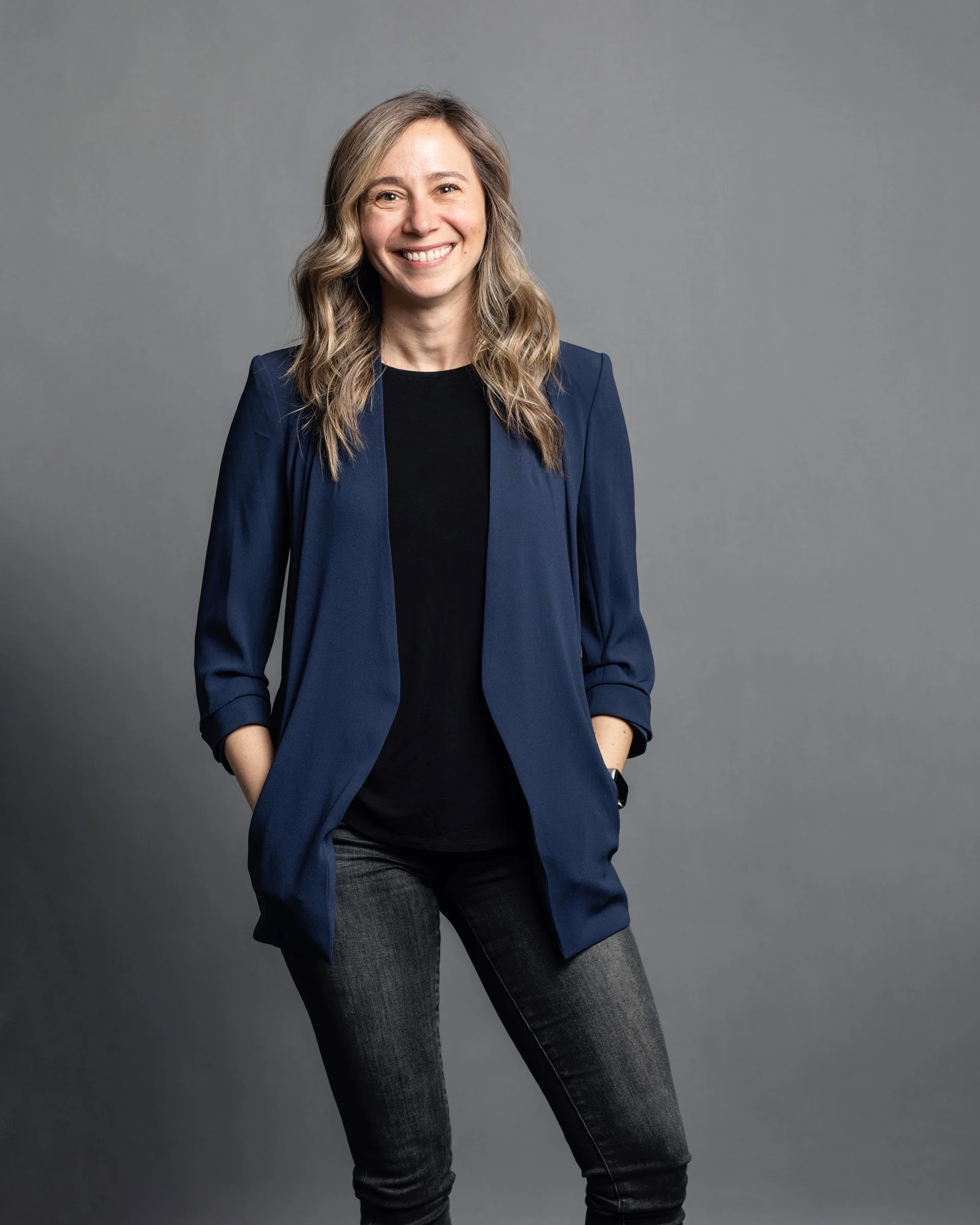 A woman with shoulder-length wavy blonde hair smiles while standing against a gray background. She wears a navy blazer over a black shirt and dark gray jeans, with her hands in her pockets.
