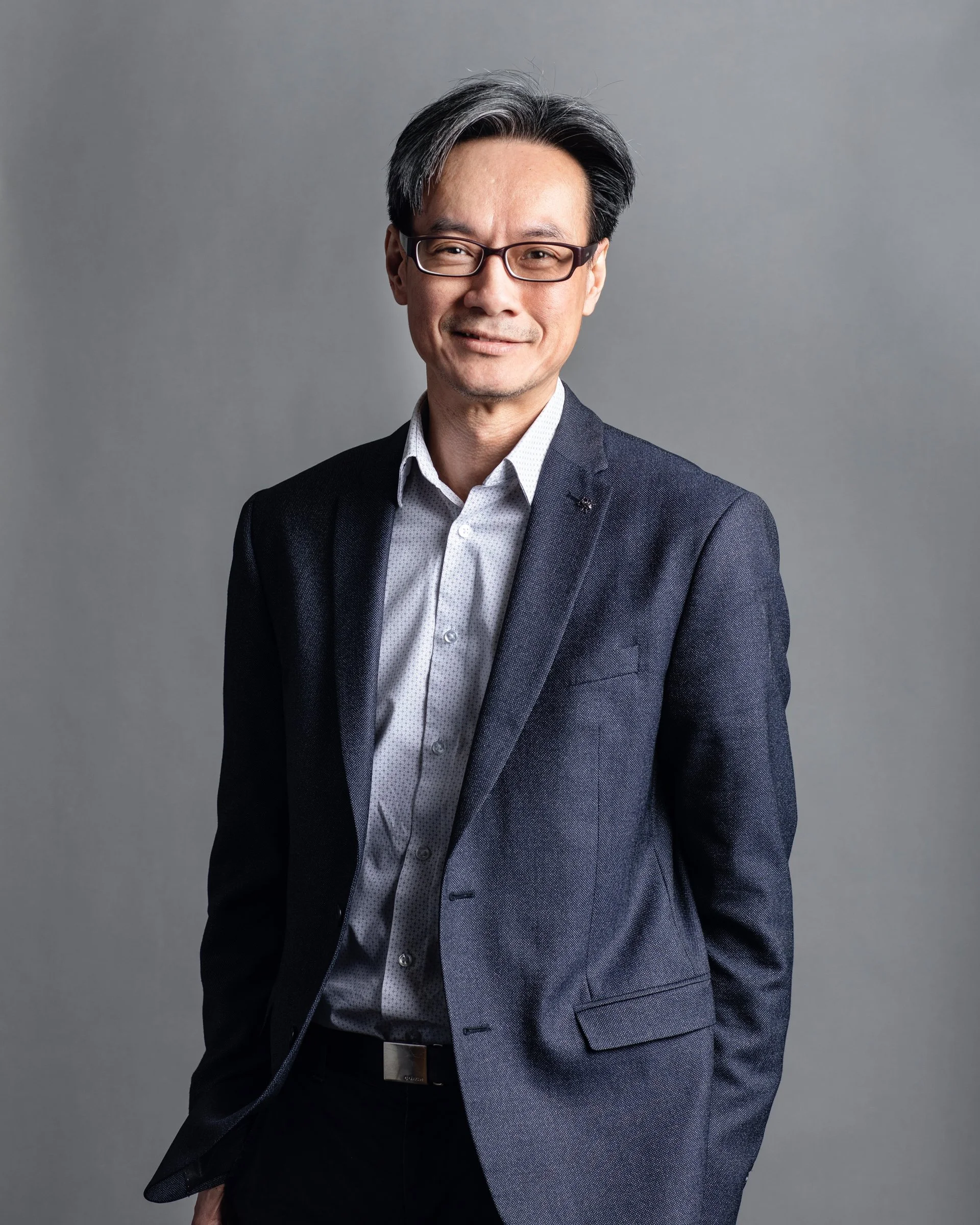 Portrait of a middle-aged man with glasses, dressed in a dark blazer and light shirt, standing against a gray background.