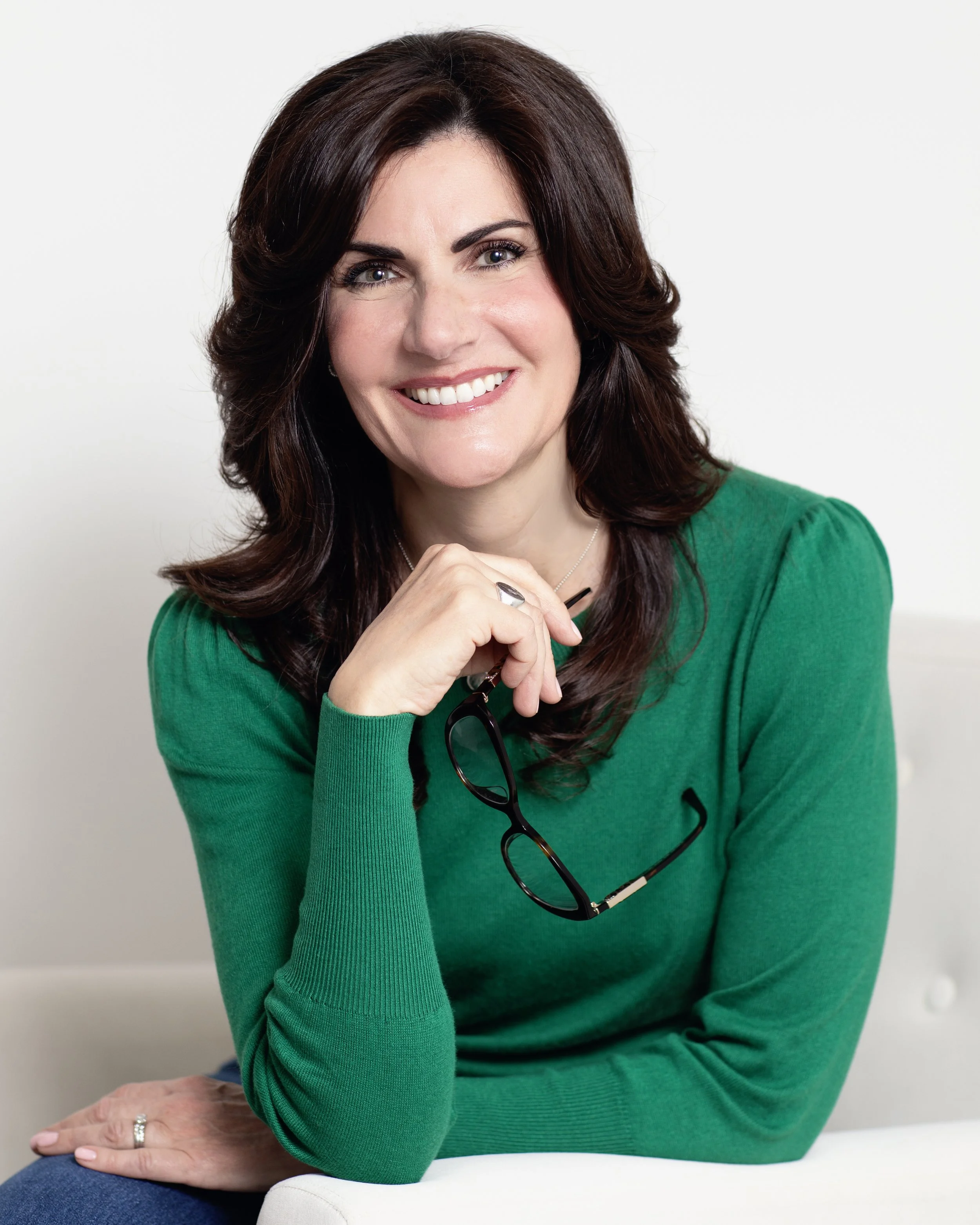 A woman with shoulder-length dark brown hair, wearing a green long-sleeve top, smiling and holding glasses in her right hand.