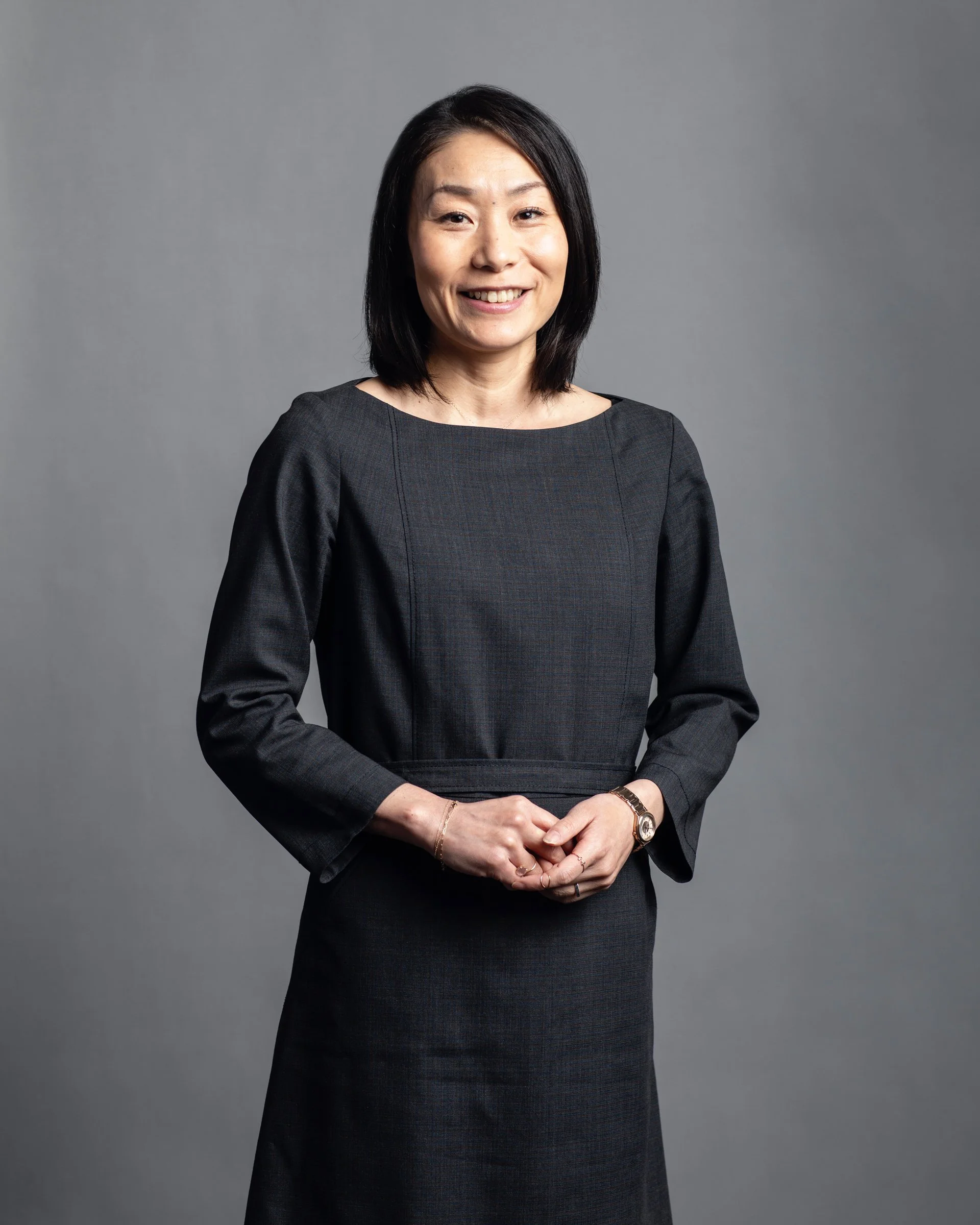 A woman with shoulder-length black hair wearing a dark dress standing against a gray backdrop, smiling, with her hands clasped in front of her.