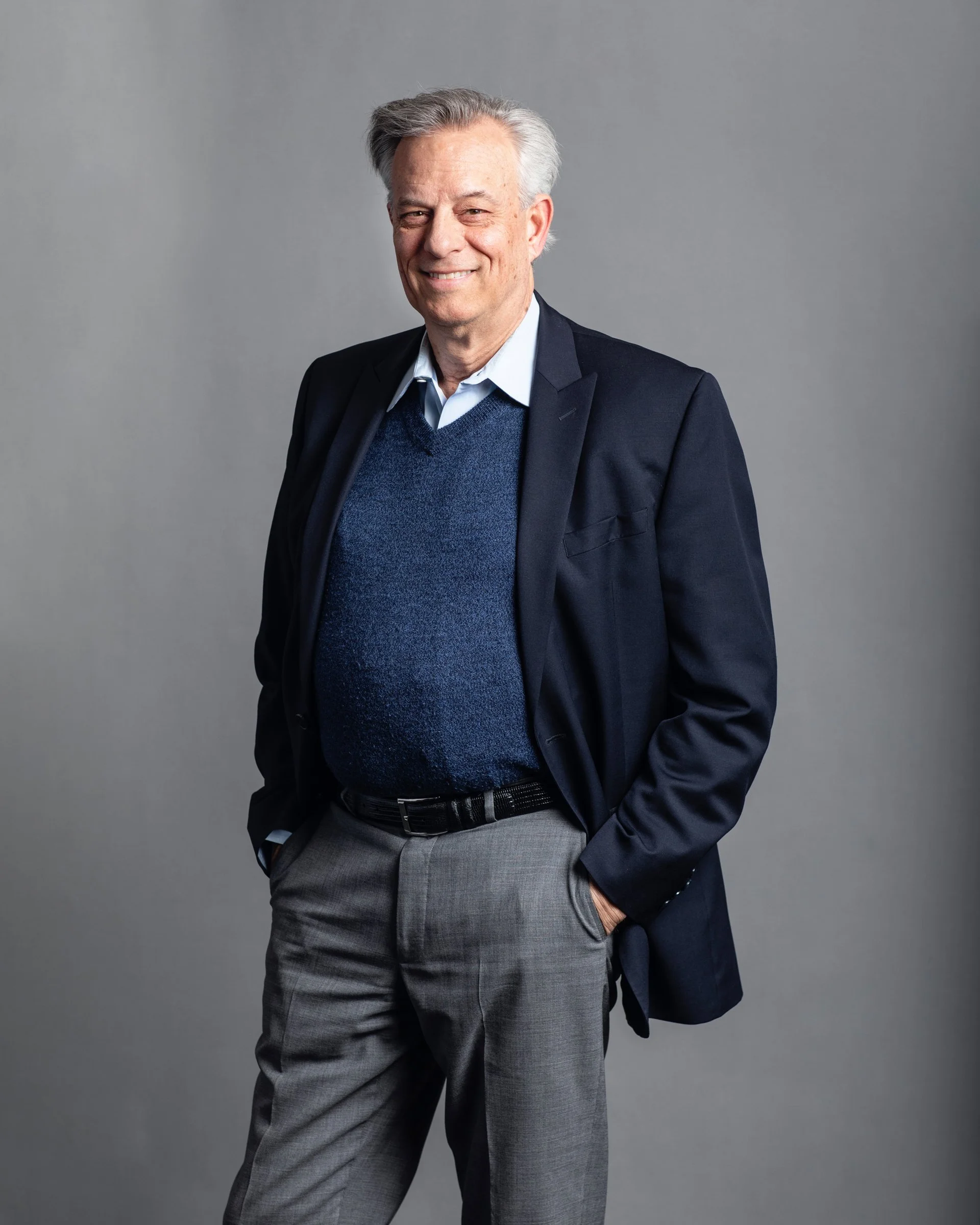 A smiling older man with gray hair, dressed in a navy blazer, light blue shirt, dark blue sweater, and gray pants, standing against a plain gray background.